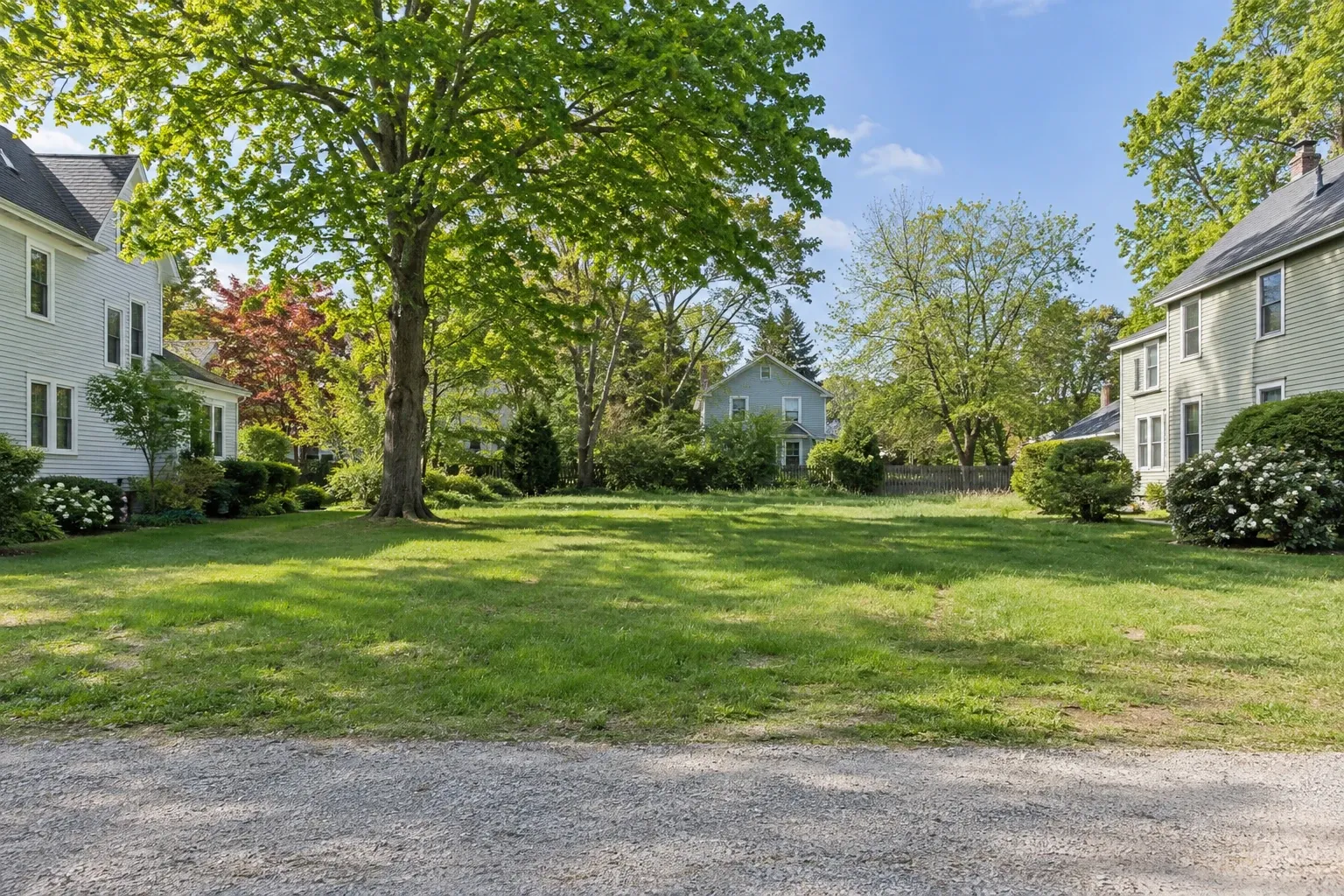 Grassy residential lot between traditional clapboard homes and mature trees