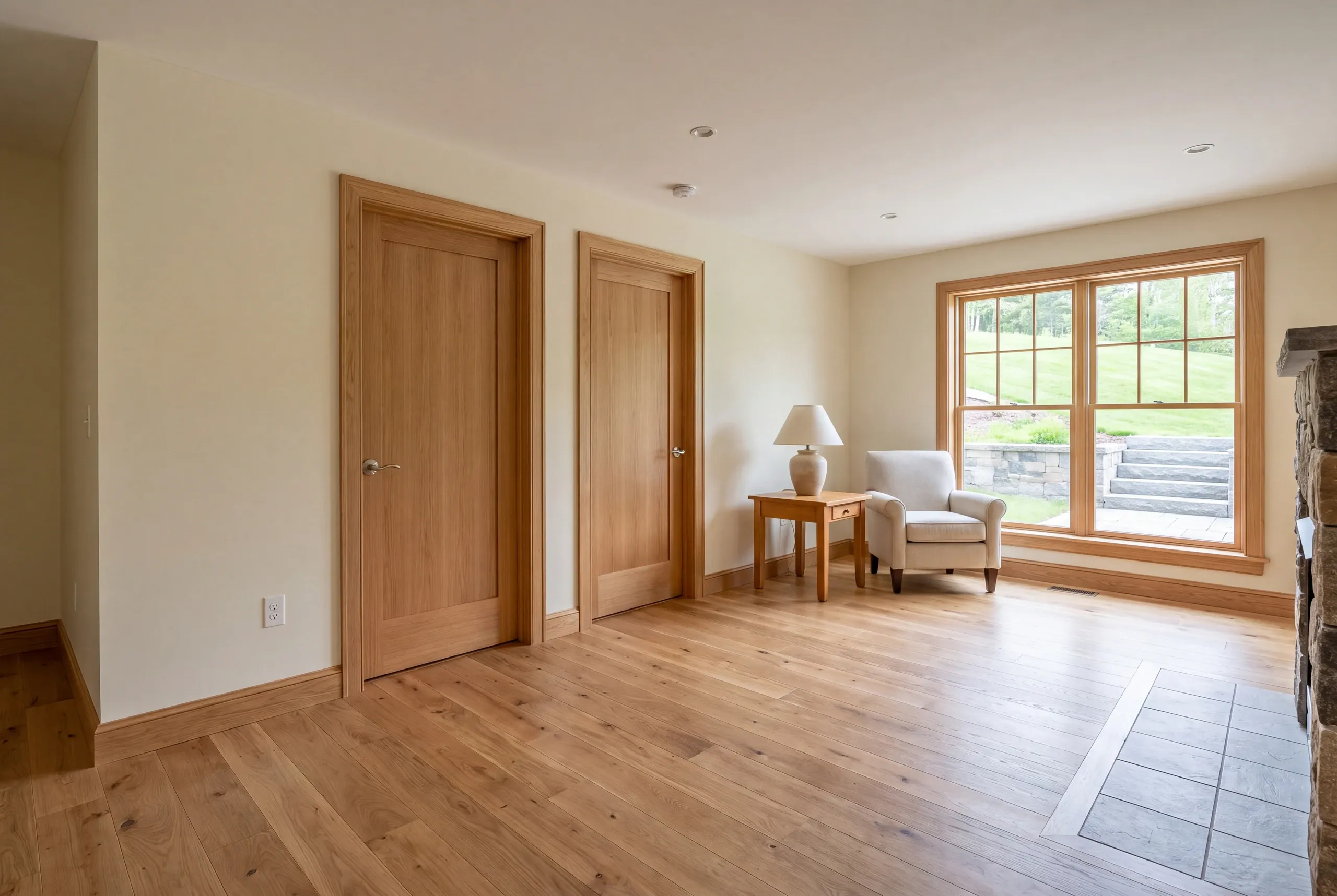 A finished lower-level room with light wood floors, two closed wooden doors, an armchair next to a small table with a lamp, and a window looking out to a grassy slope.