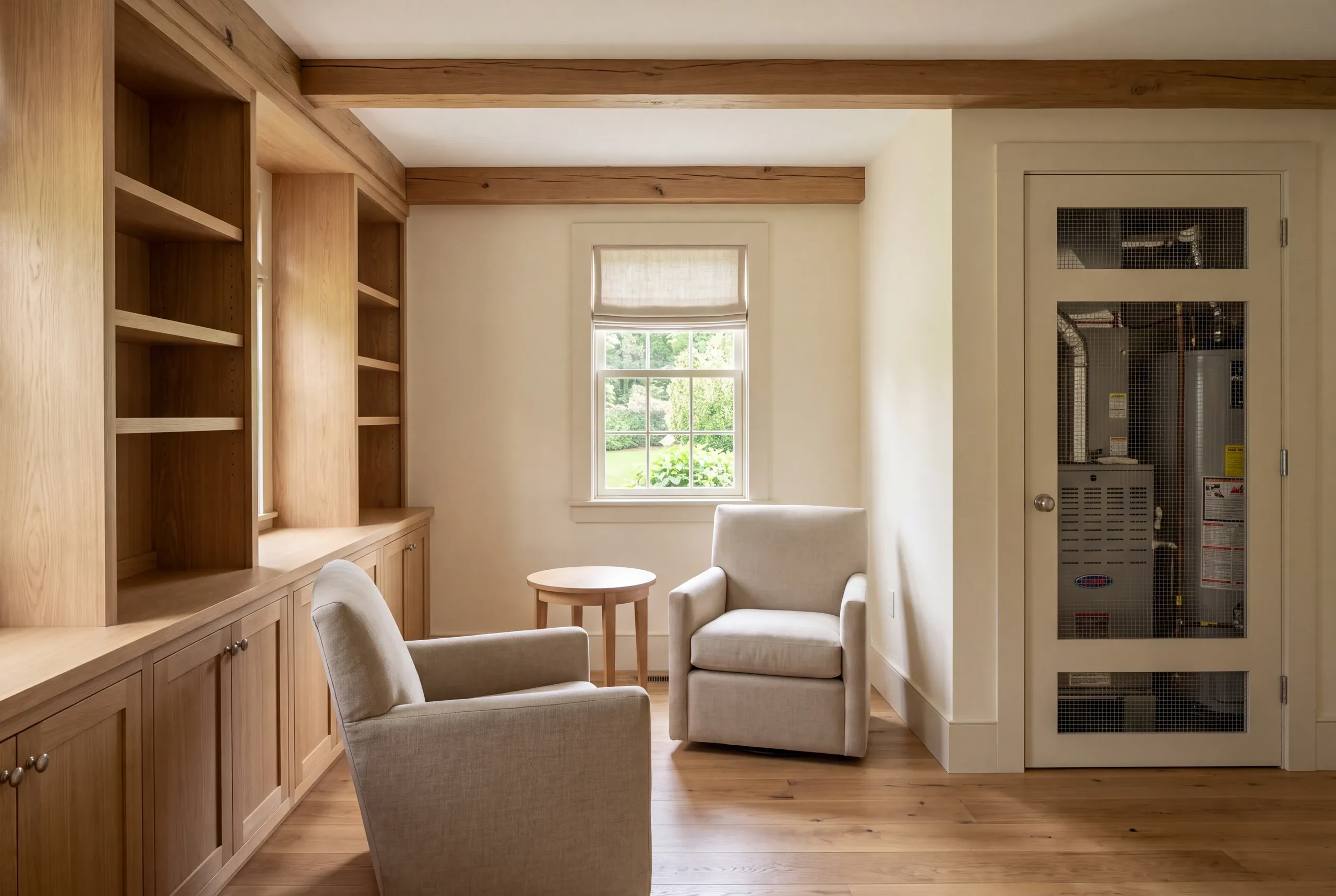 Basement remodeling in Arrowsic, ME: interior room featuring built-in wooden shelving, two armchairs, a small round table, and a door with wire mesh panels showing utility equipment inside.