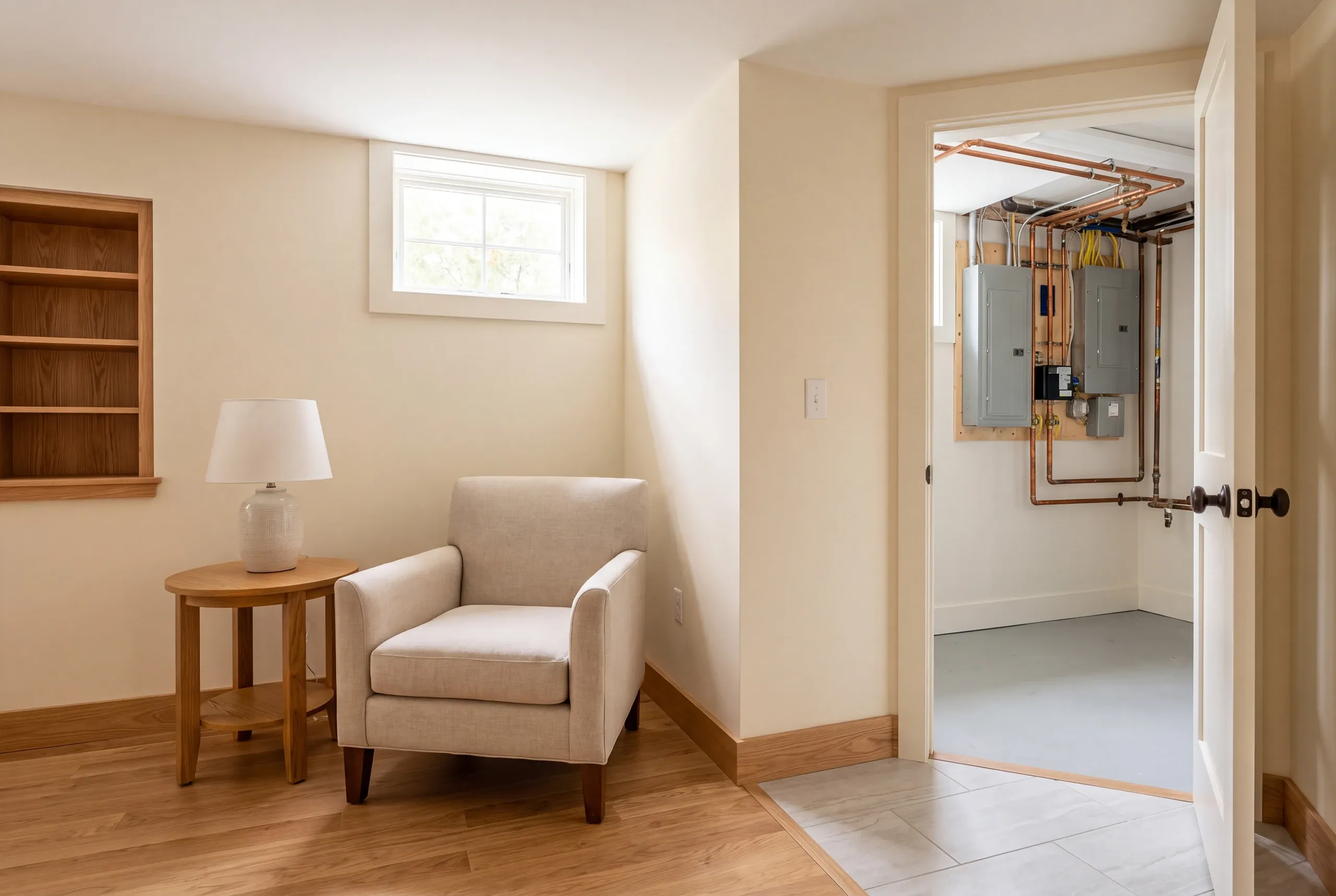 Basement remodeling in Bath, ME: a finished basement room with beige walls, an armchair, a side table with a lamp, built-in shelving, and an open doorway revealing utility electrical panels and copper plumbing.