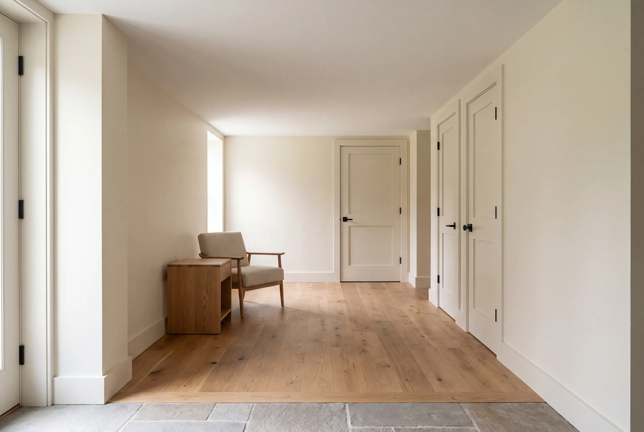 A finished hallway with light wood flooring, beige walls, several closed white doors, and a small chair with a wooden side table.