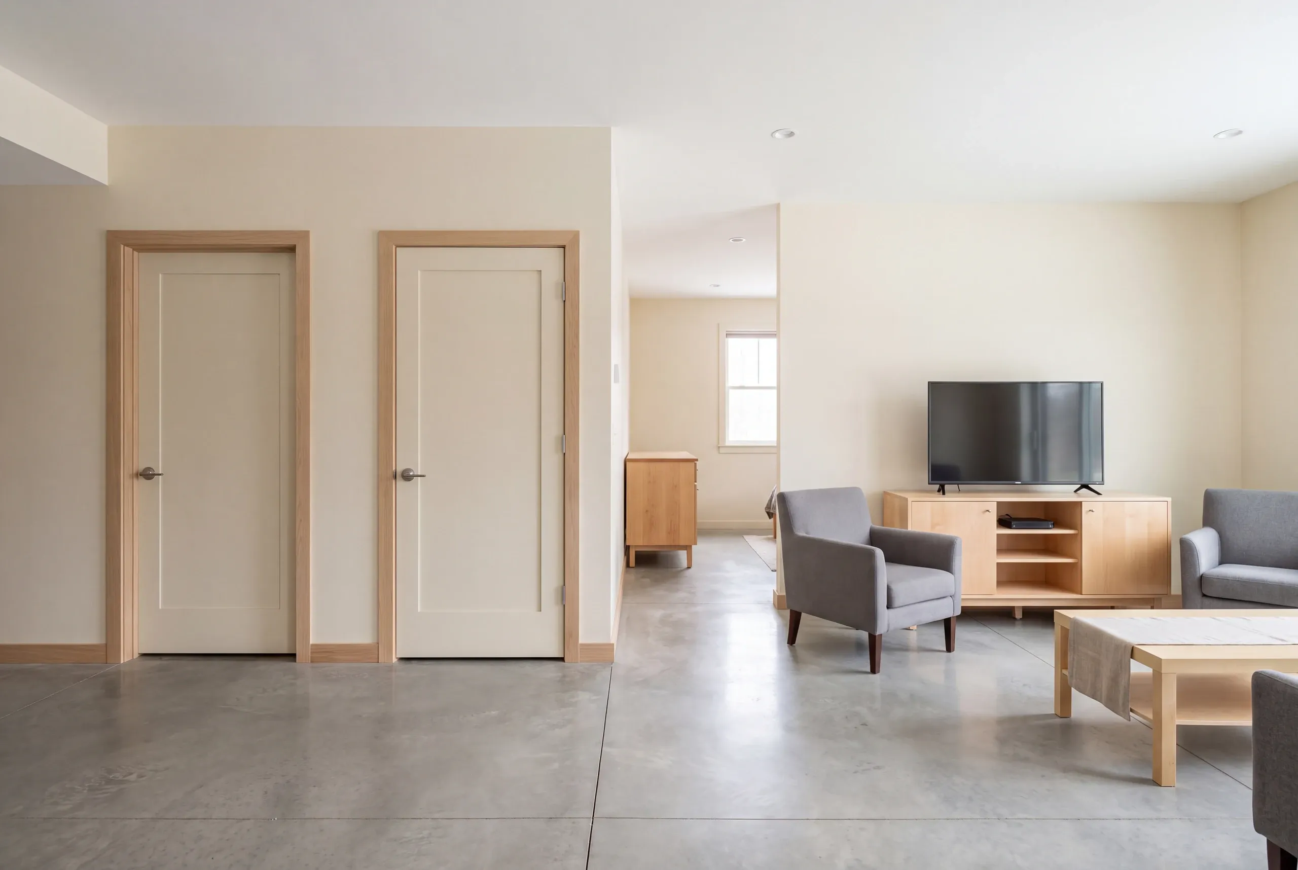 A finished lower level recreation room with concrete floors, two closed doors on the left, and a seating area with a television on the right.