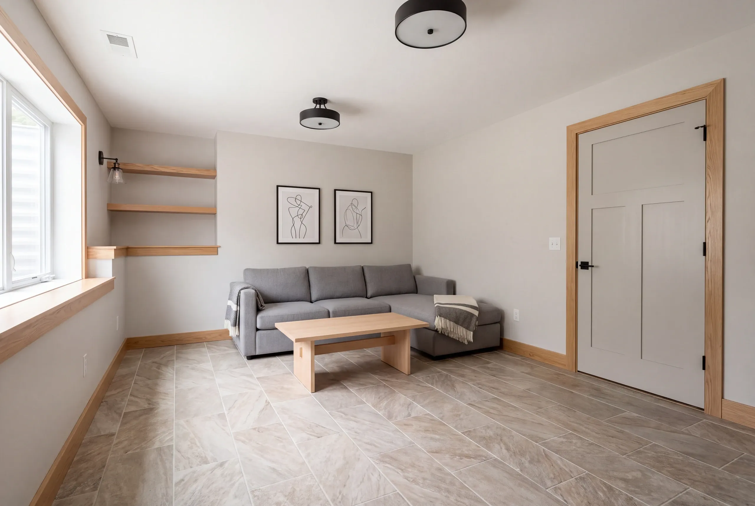 Interior shot of a finished recreation room with a gray sectional sofa, wooden coffee table, tile flooring, and light wood trim.
