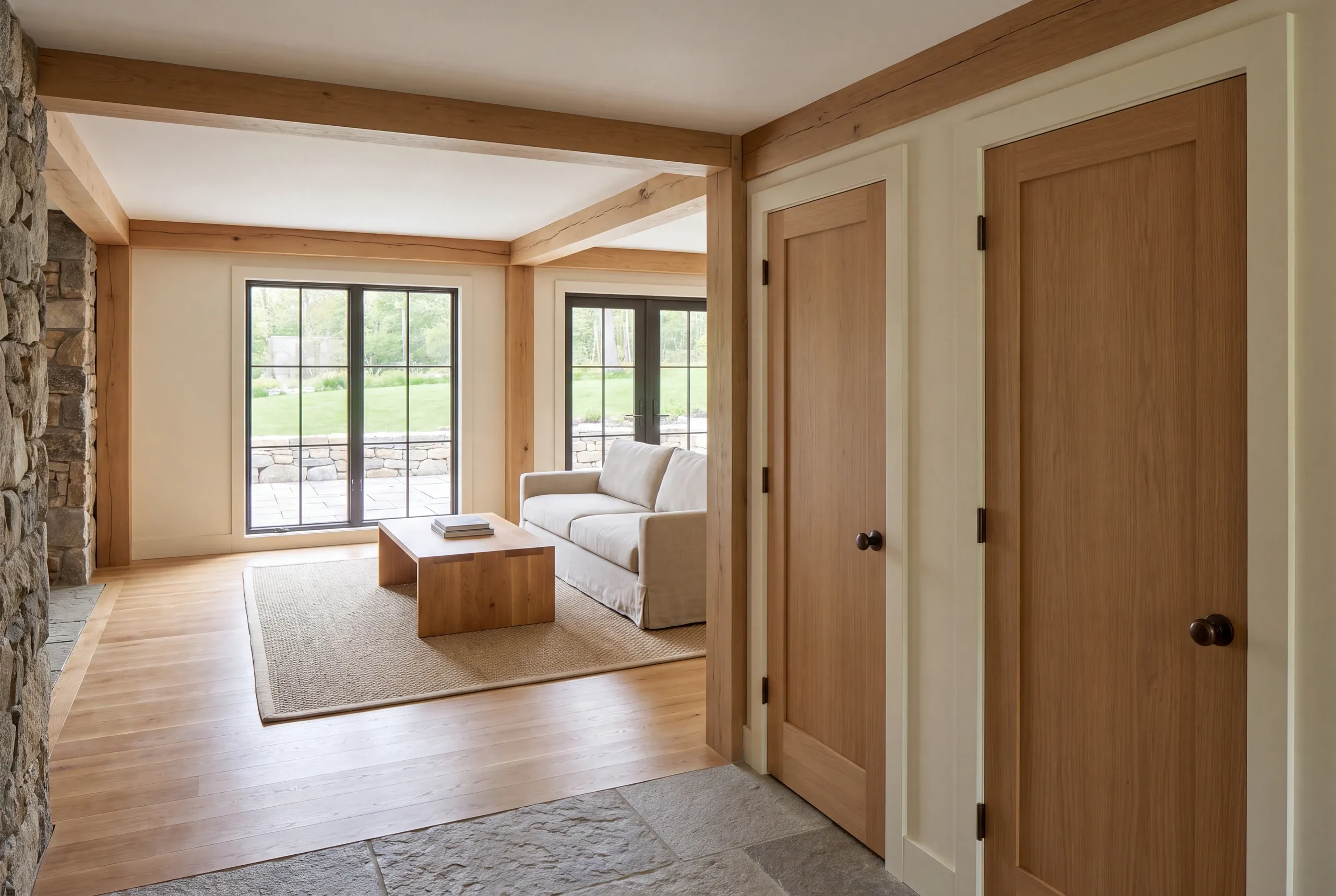 Interior view showing a finished room with exposed wooden beams, hardwood floors, closed wooden doors on the right, and a seating area with a sofa and coffee table near large windows looking out to a green lawn.