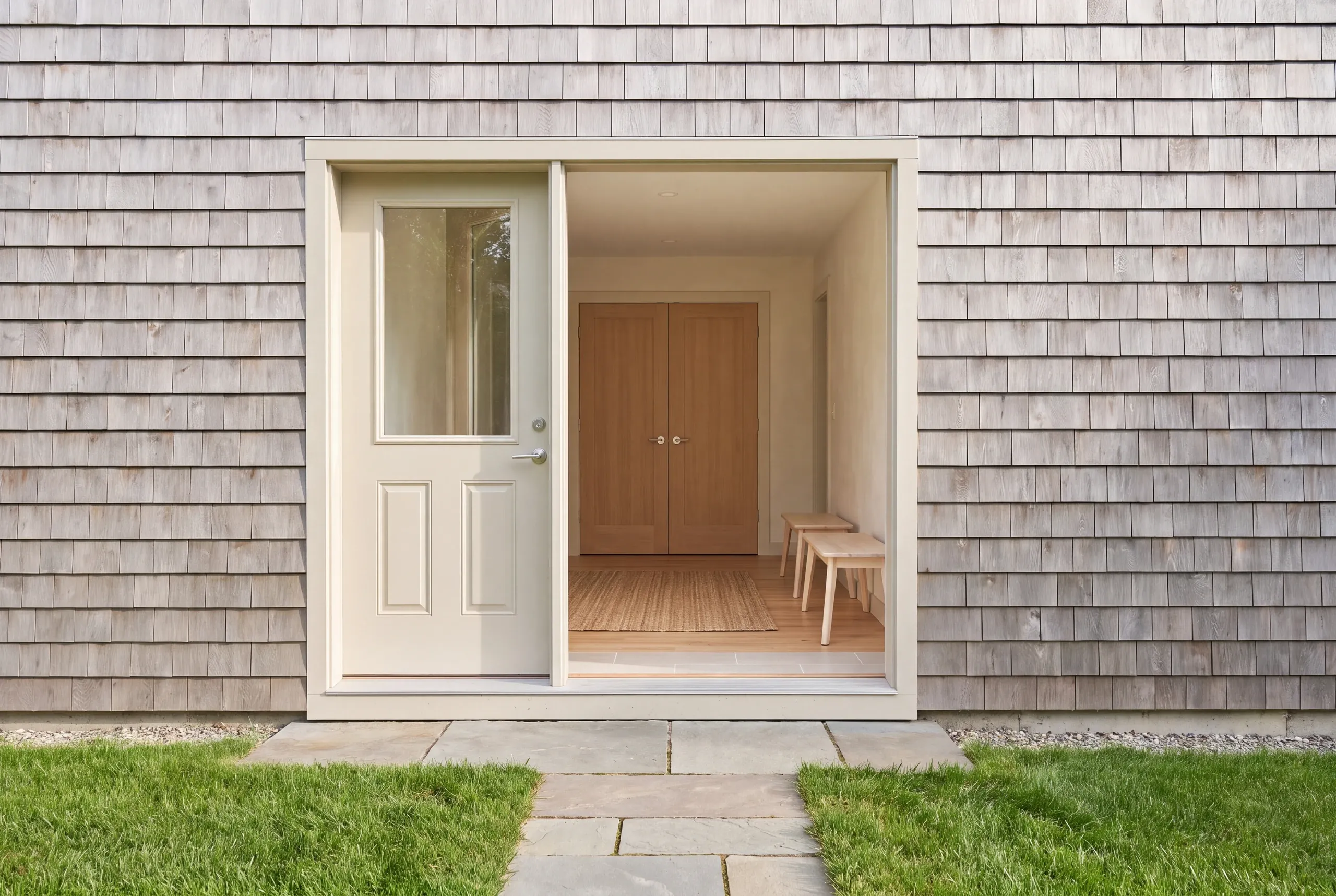 An exterior view showing a light-colored wooden shingle facade with an open doorway revealing an interior room and closed double doors.
