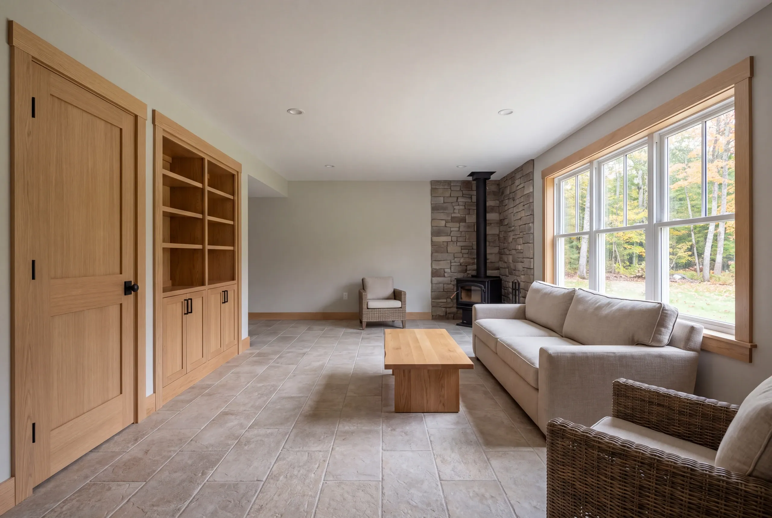 A finished lower-level recreation room featuring a tiled floor, light walls, wooden built-in shelving, a sofa, chairs, and a wood stove against a stone accent wall.