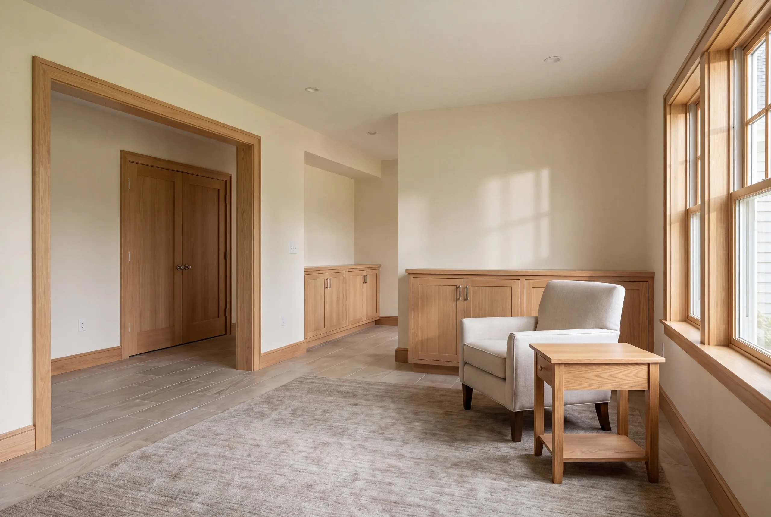 A finished room features wood trim, built-in cabinetry, a light-colored armchair, a side table, and closed wooden doors in the background.
