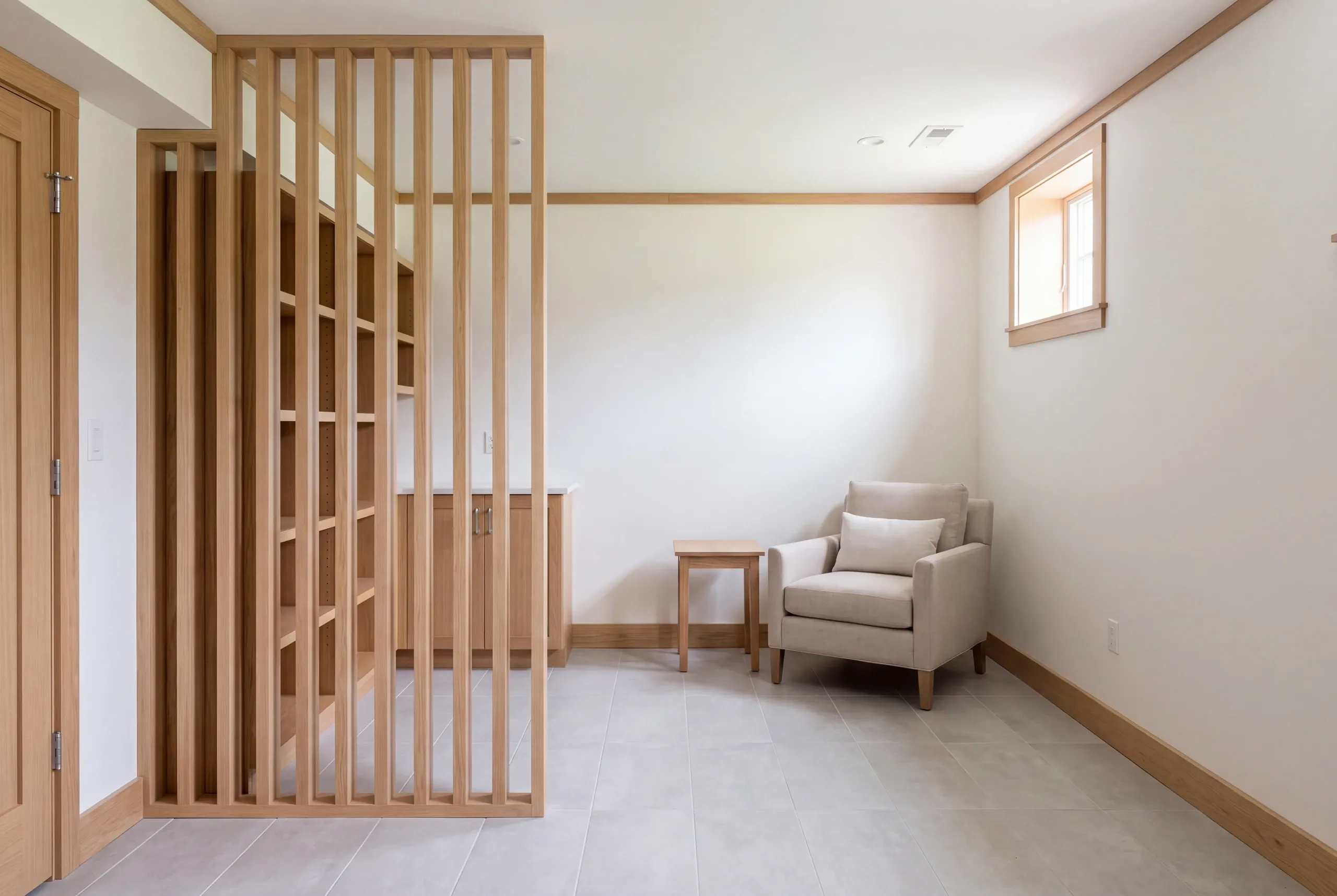 A newly finished basement room featuring a wooden slatted partition separating a shelving and storage area from a sitting space with an armchair and small side table.