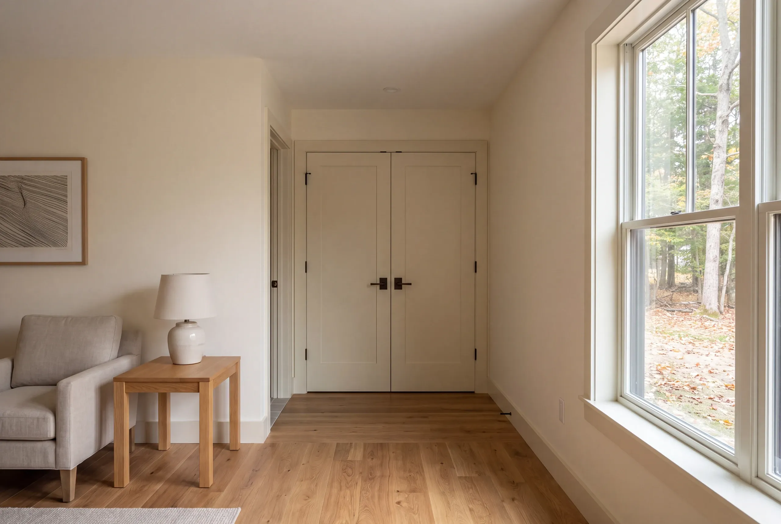 A finished interior room with light beige walls, hardwood floors, a closed set of double doors, a side table with a lamp, an armchair, and a large window looking out onto trees.