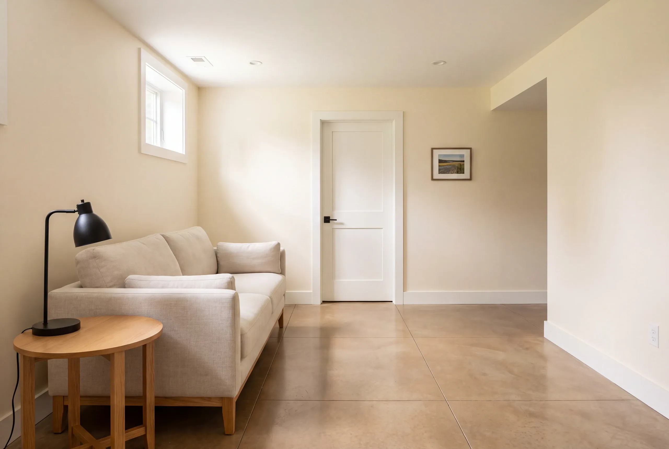 A finished basement room features a beige sofa, a small wooden side table with a black lamp, tiled floors, and a closed white interior door.