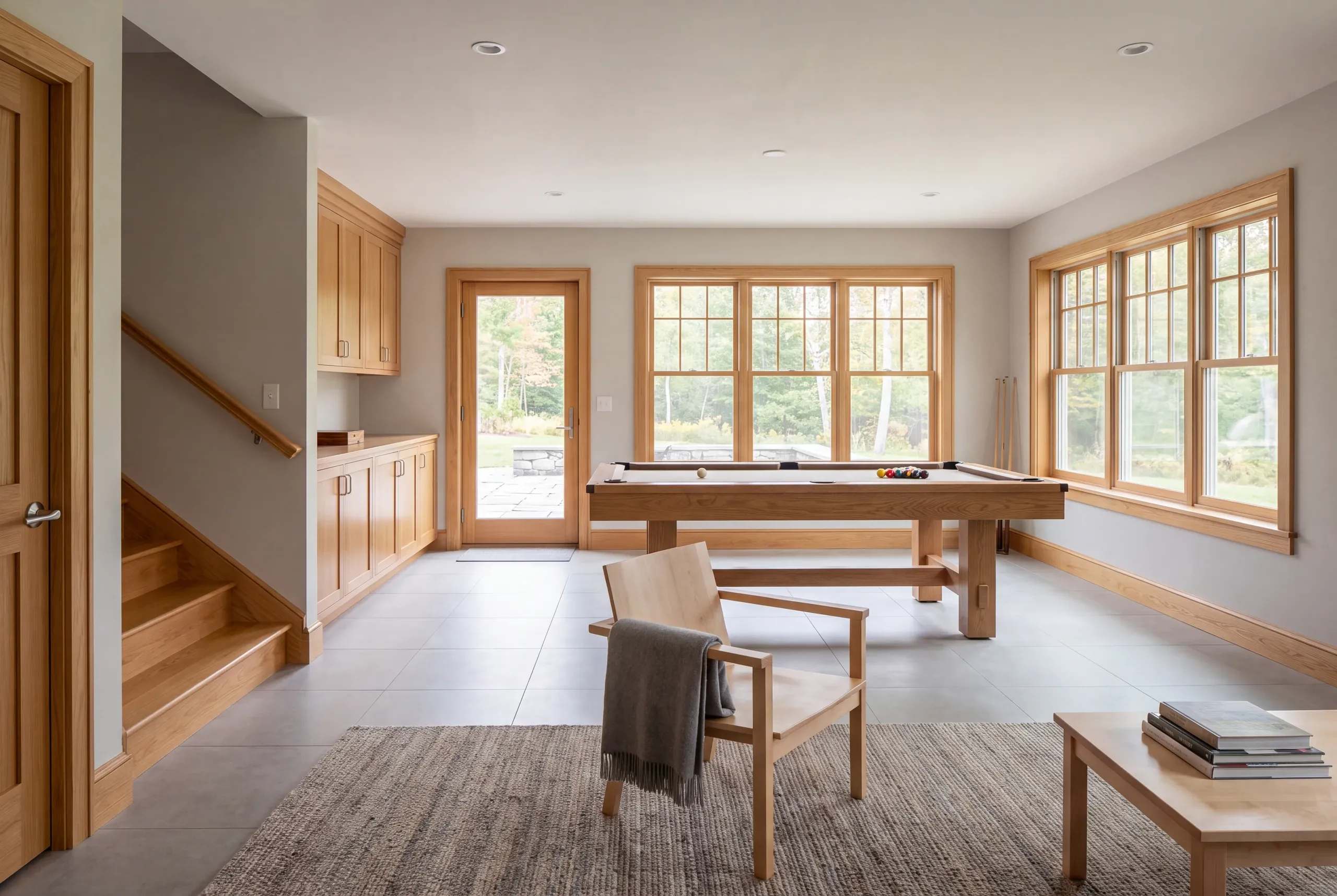 A finished lower-level recreation room features a wooden pool table, light wood trim, a staircase, cabinetry, and a seating area with a wooden armchair.