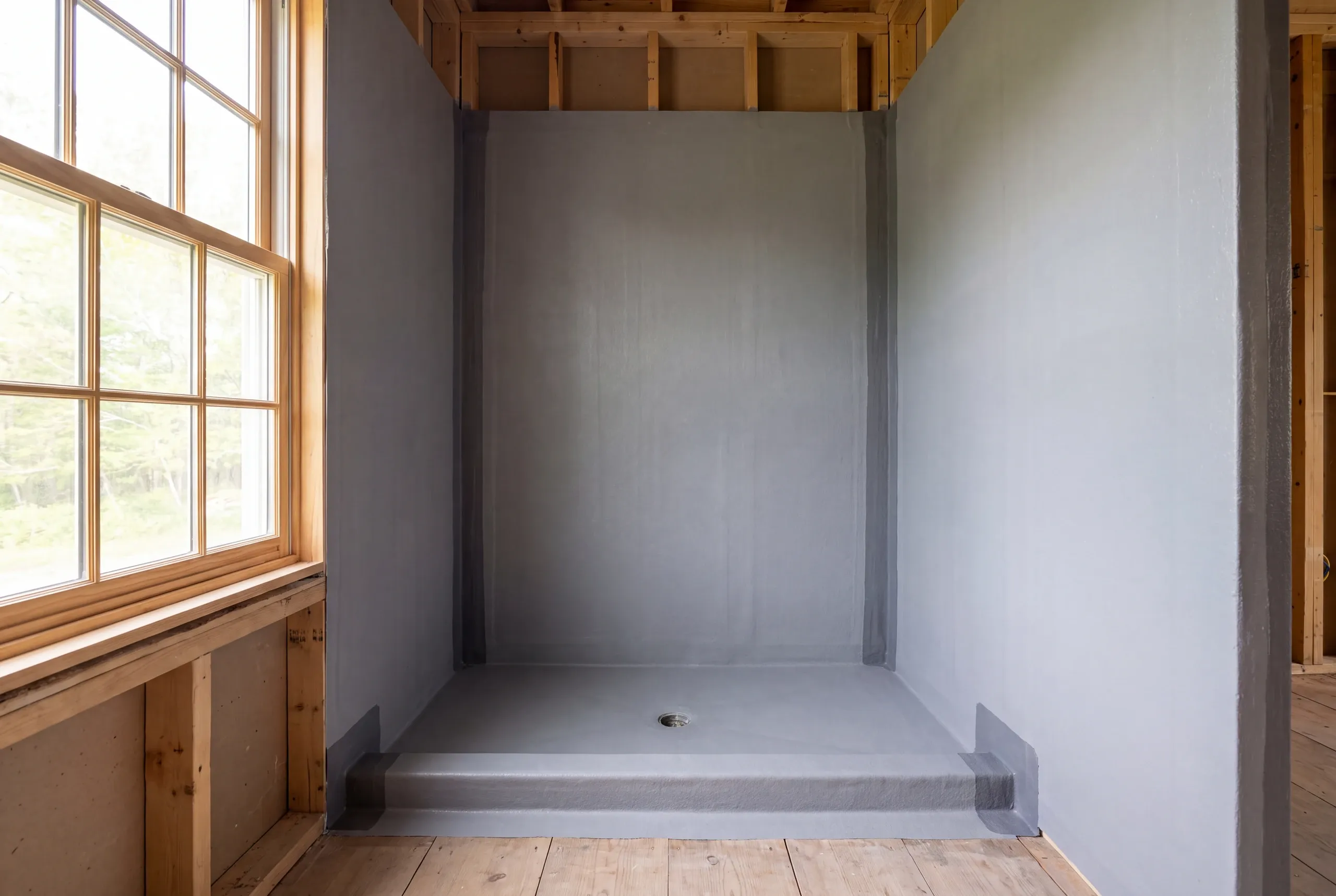 An empty shower alcove with grey waterproofing membrane applied to the walls, pan, and curb, alongside exposed wood framing and a window.