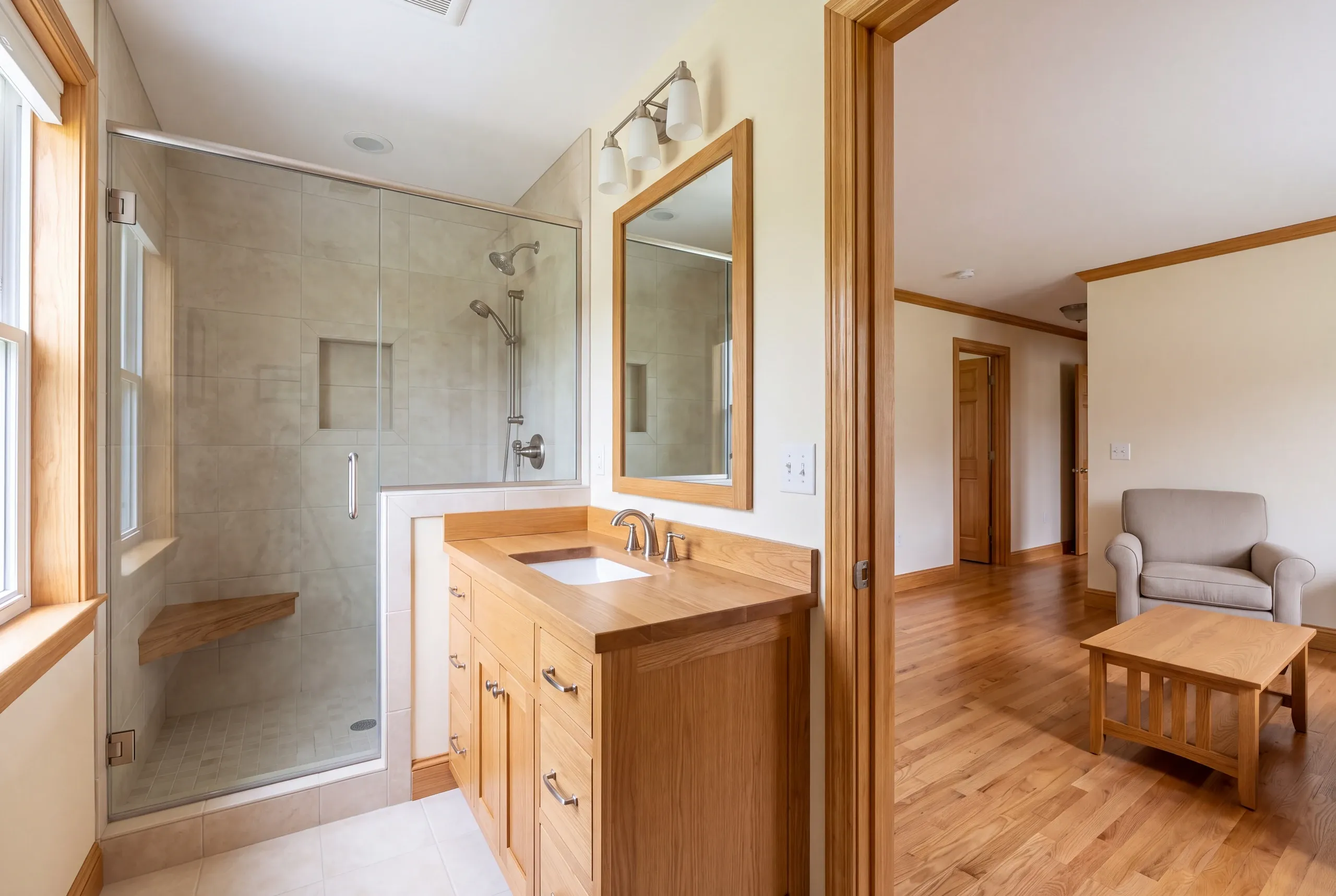 A finished bathroom features a glass-enclosed tile shower with a wooden bench, a wooden vanity with a sink and mirror, and an open doorway leading to a hardwood-floored room with furniture.