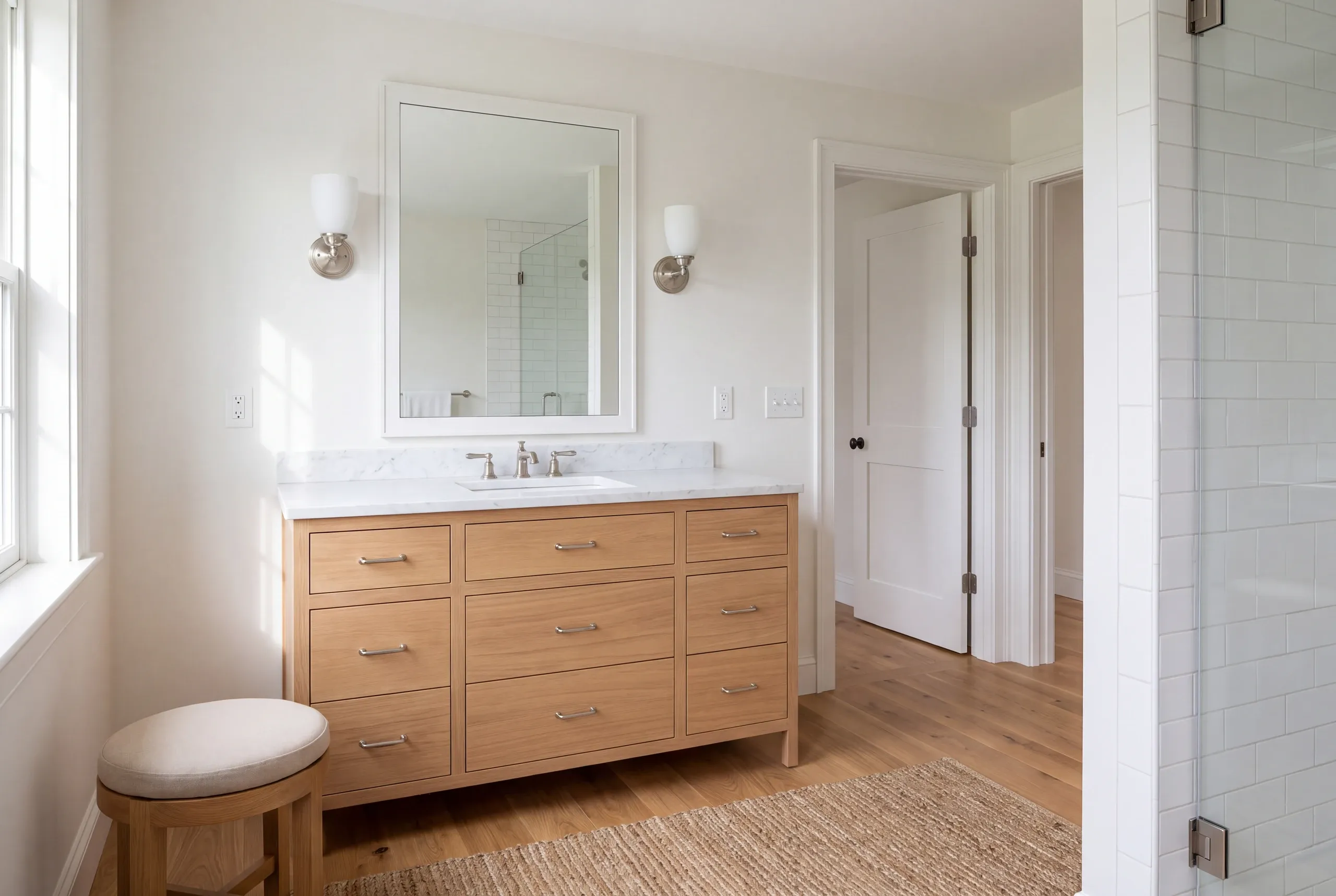 A finished bathroom with a wooden vanity, marble countertop, mirror, sconces, a small stool, and an adjacent walk-in shower.