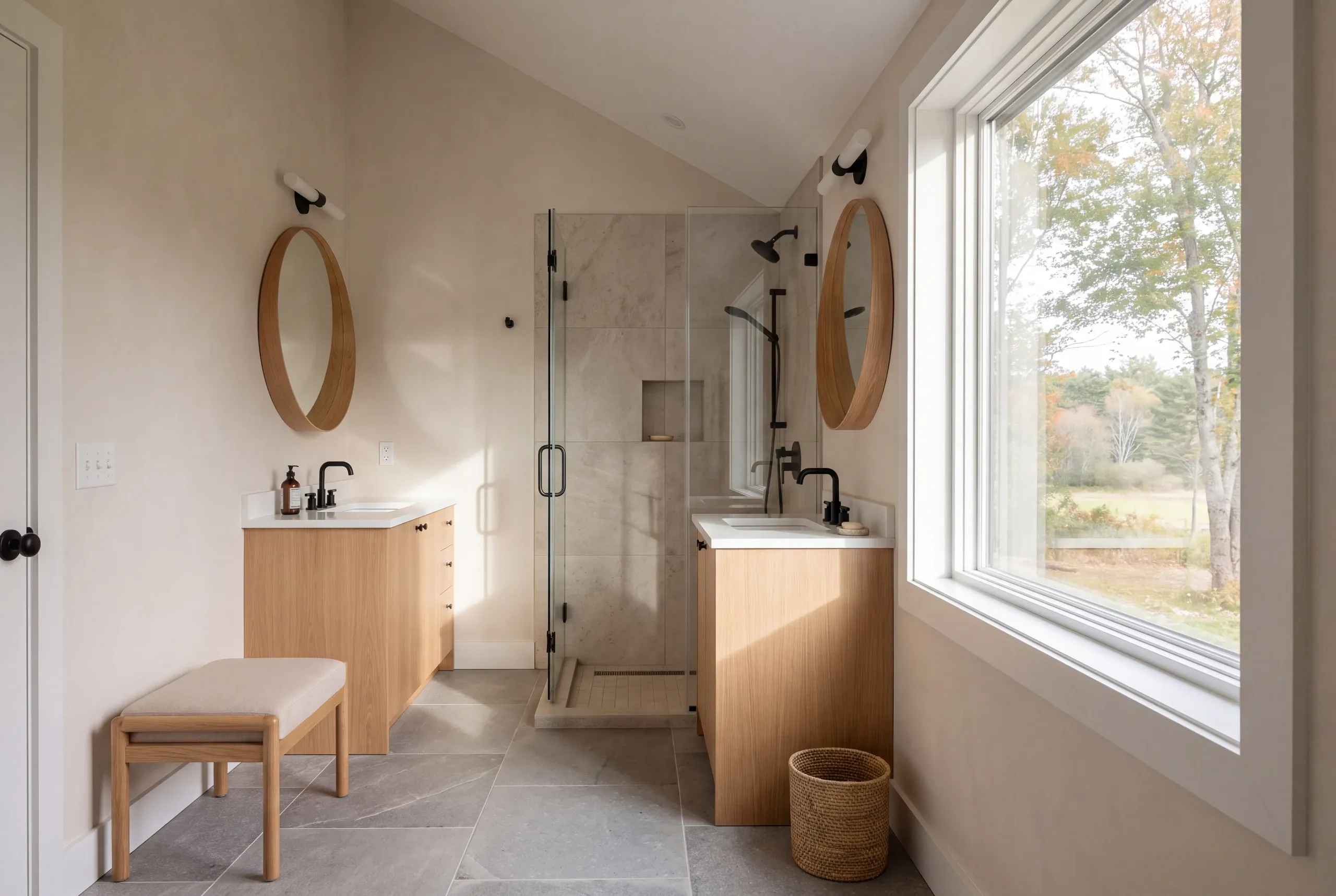 A bright bathroom features two wooden vanities with round mirrors, a glass-enclosed shower, and a large window overlooking autumn trees.