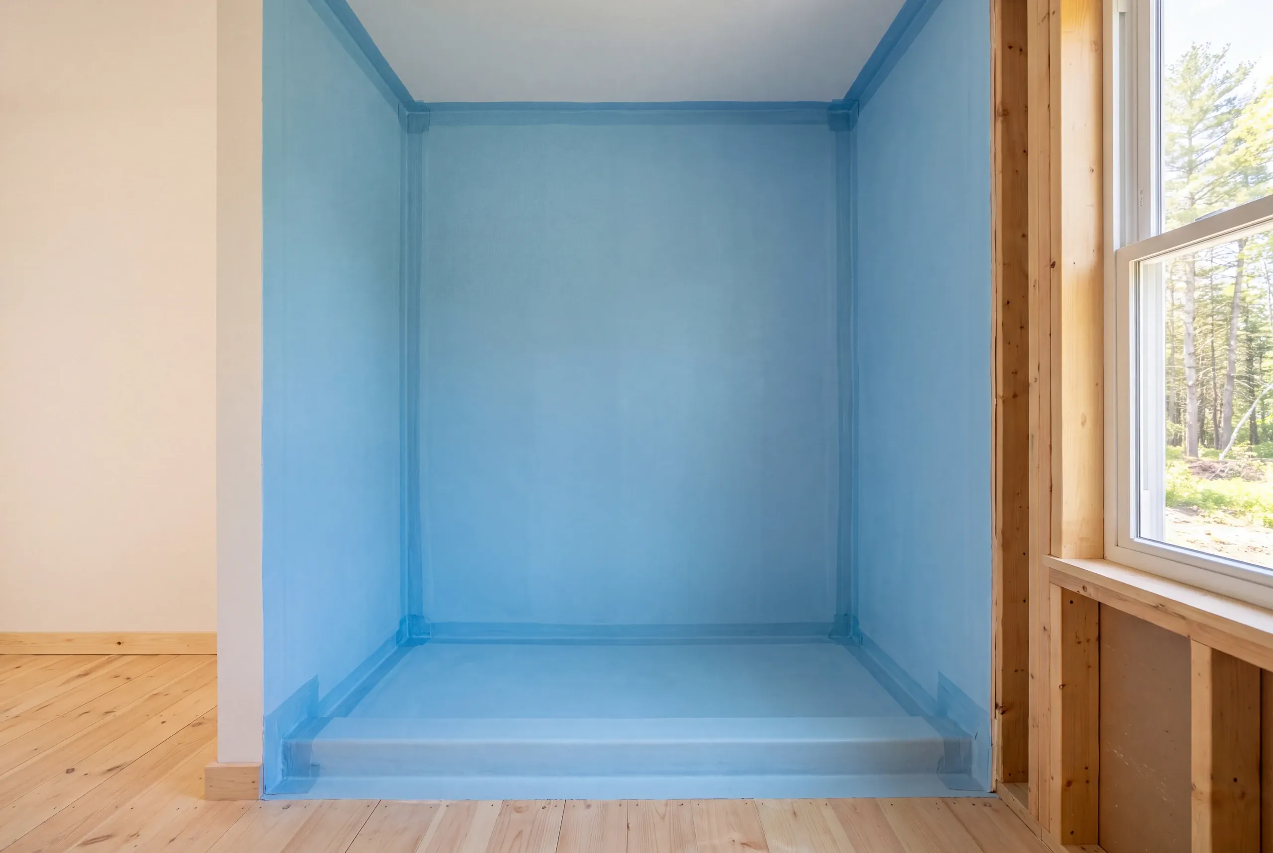An empty shower alcove with blue waterproof membrane applied to the walls, floor pan, and sealed corners, with exposed wood framing to the right and a window looking out at trees.