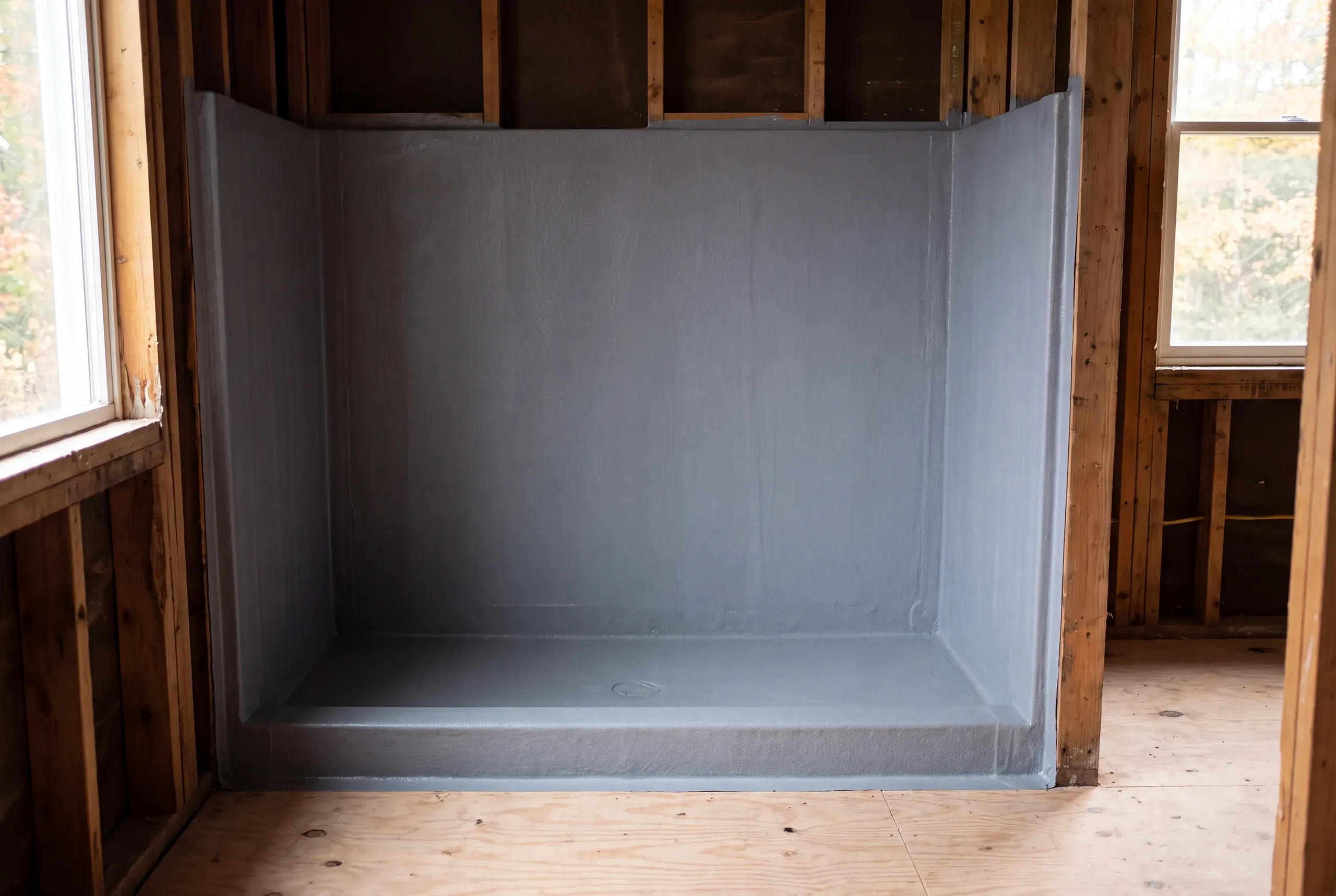 An empty shower alcove coated with a gray continuous waterproofing membrane in a room with exposed wood framing and plywood subflooring.