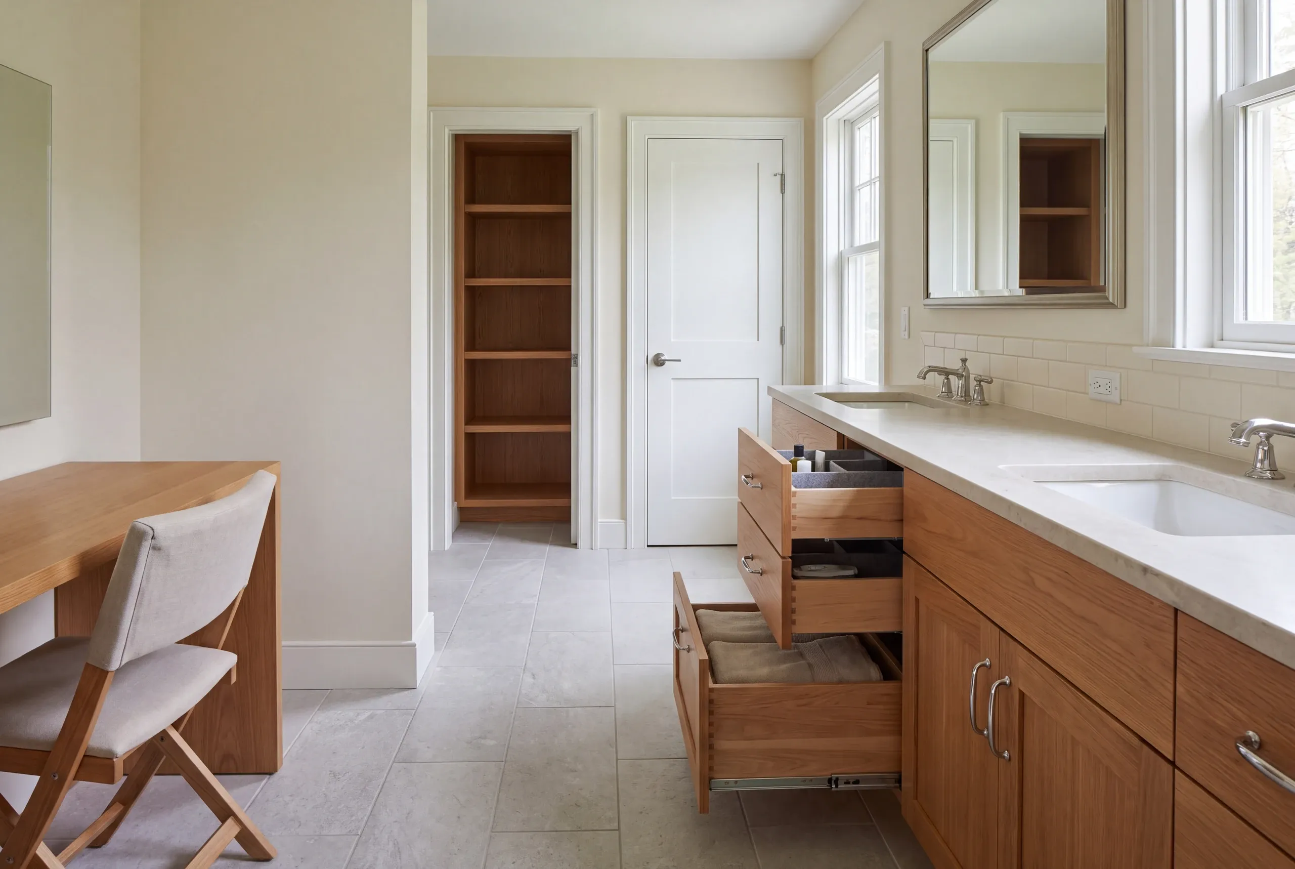 A brightly lit bathroom features wooden vanity cabinetry with open drawers displaying towels and organizers, alongside a matching wooden desk area and a tall open shelving unit in the background.