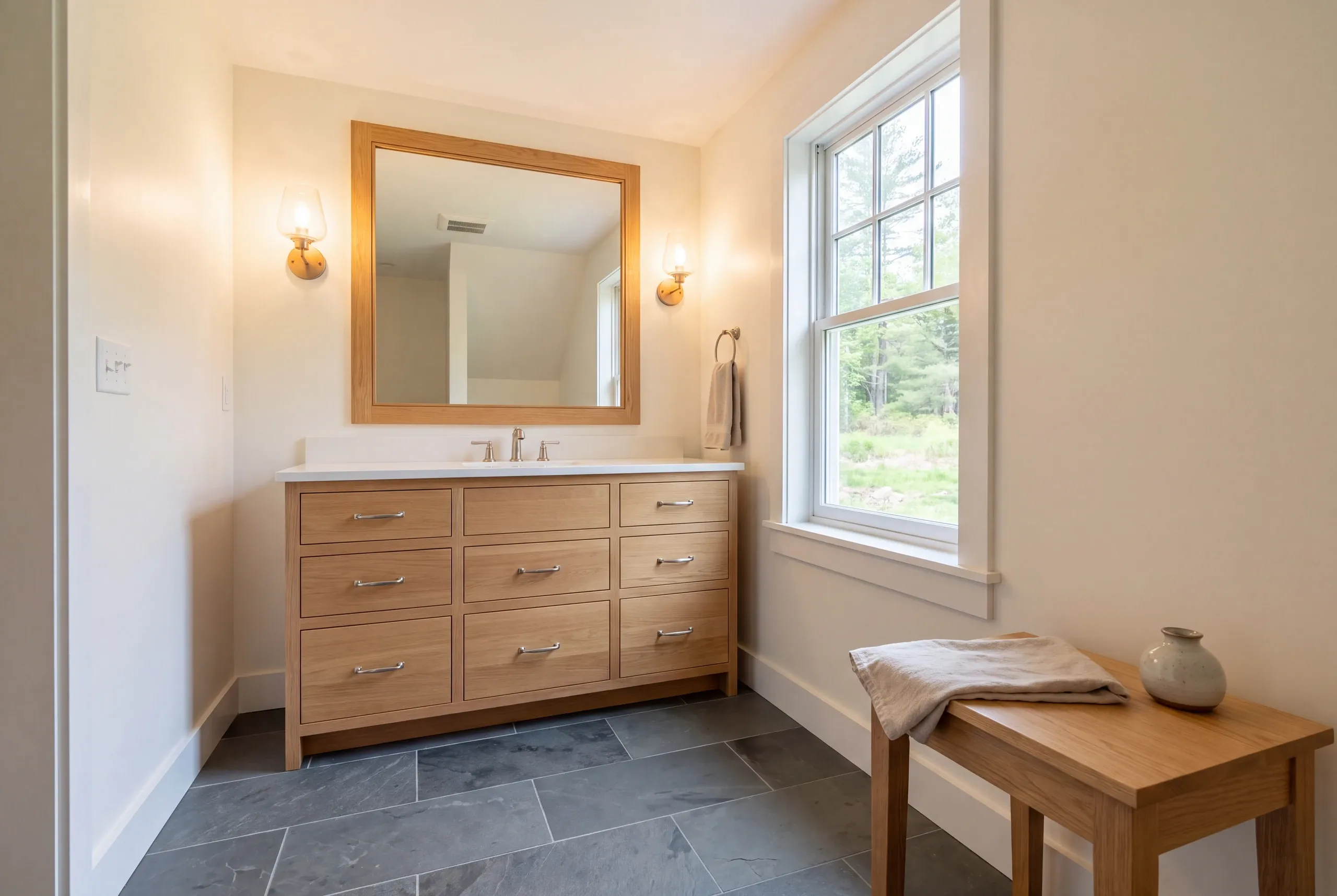 A finished bathroom features a wooden vanity with multiple drawers, a large mirror, two wall sconces, a window, dark tile flooring, and a wooden bench holding a towel and a small vase.