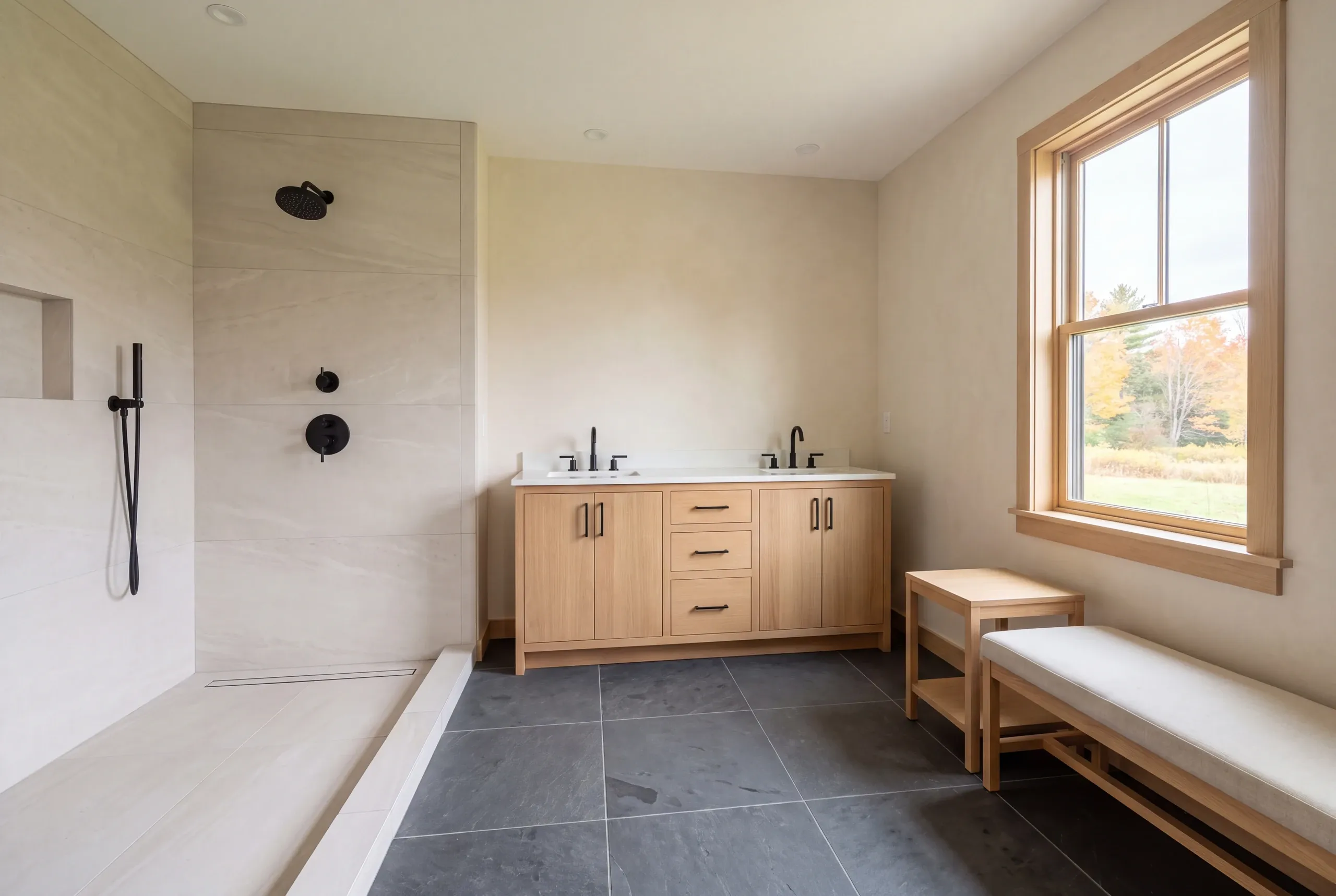 A remodeled bathroom featuring a tiled walk-in shower, a wooden double-sink vanity, and a window with a bench below.
