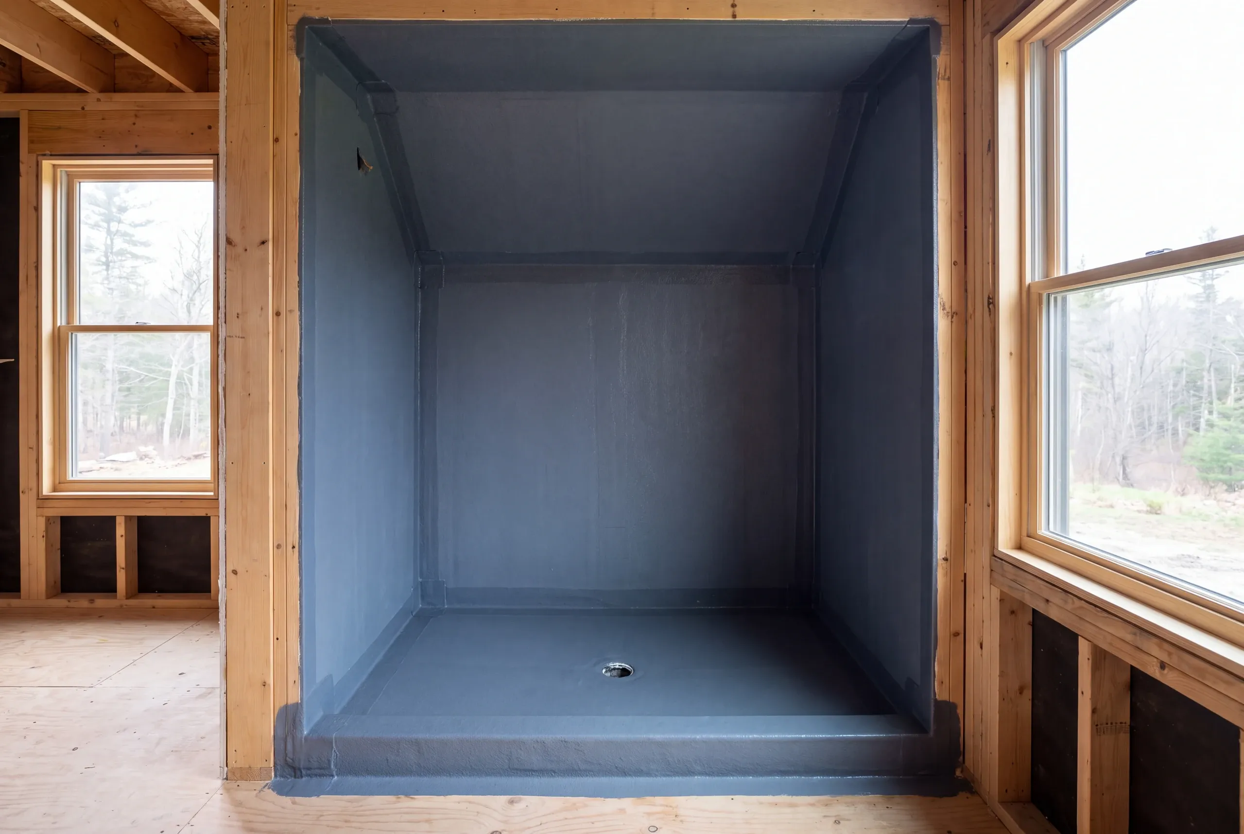 An empty shower alcove in a wood-framed room is coated with a grey continuous waterproofing membrane.