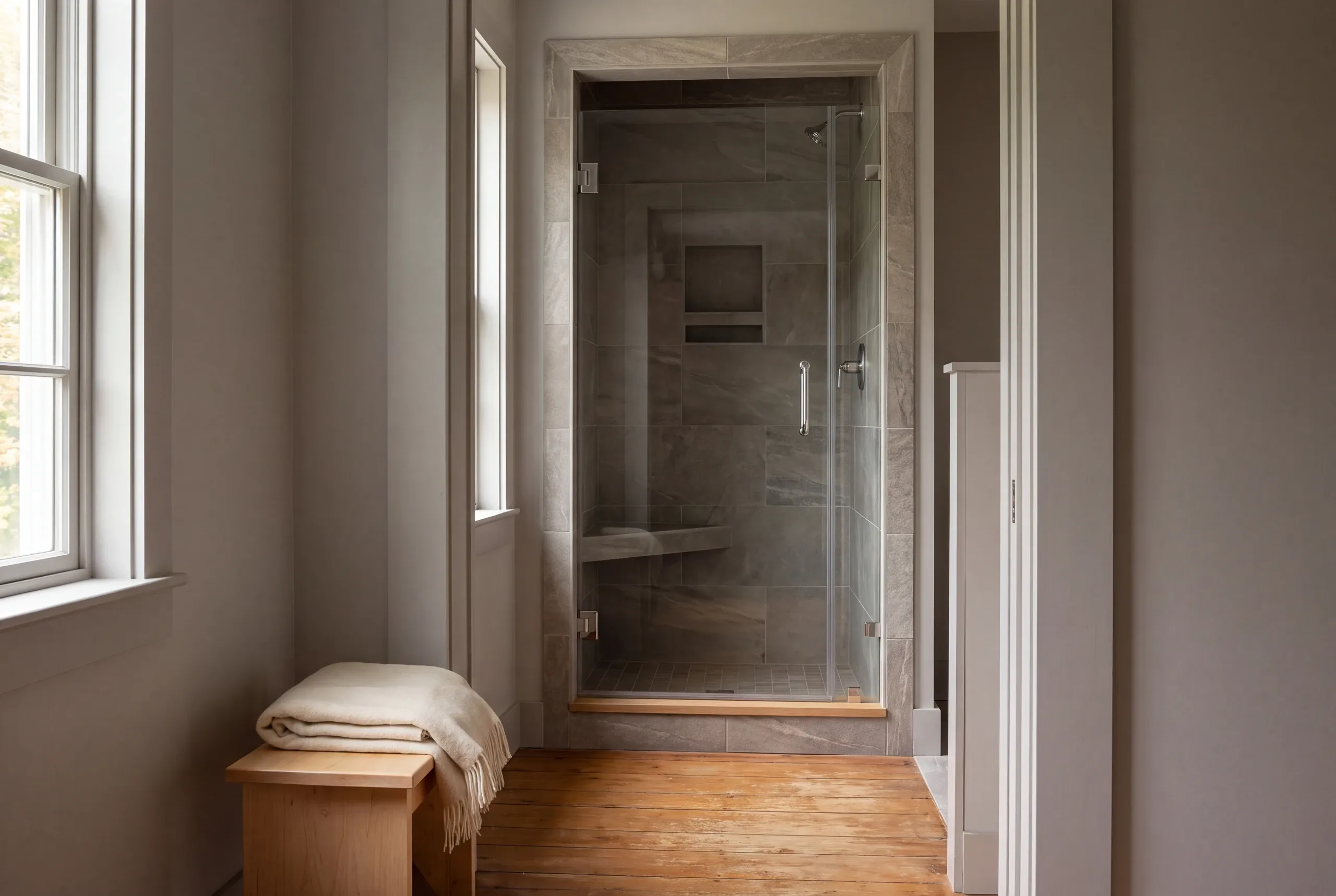 A finished tiled shower enclosure with a glass door and bench seat stands in a room with a wooden floor and a small bench holding a folded blanket.