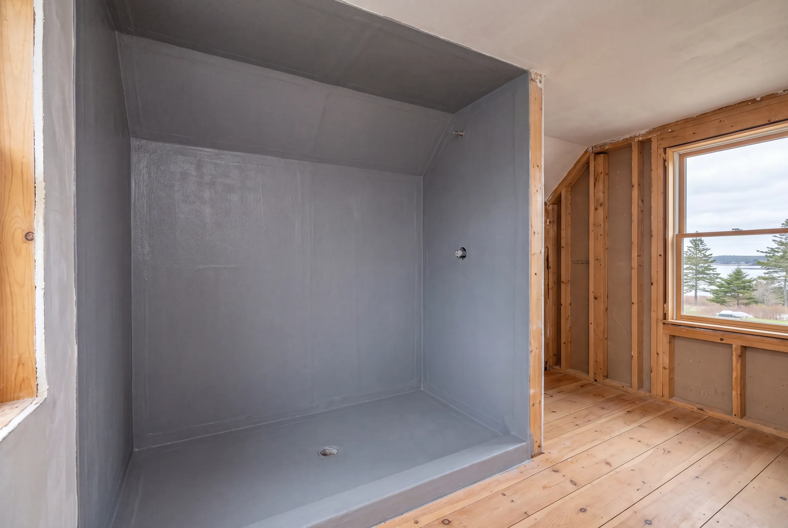 An empty shower enclosure coated in grey waterproofing material with a drain and exposed wood framing nearby.