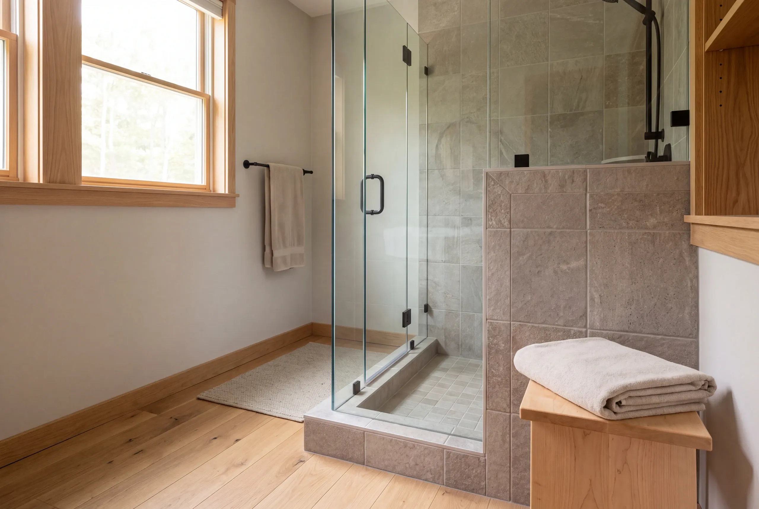 A finished tiled shower enclosure with a glass door and wooden trim stands in a bathroom with hardwood floors.