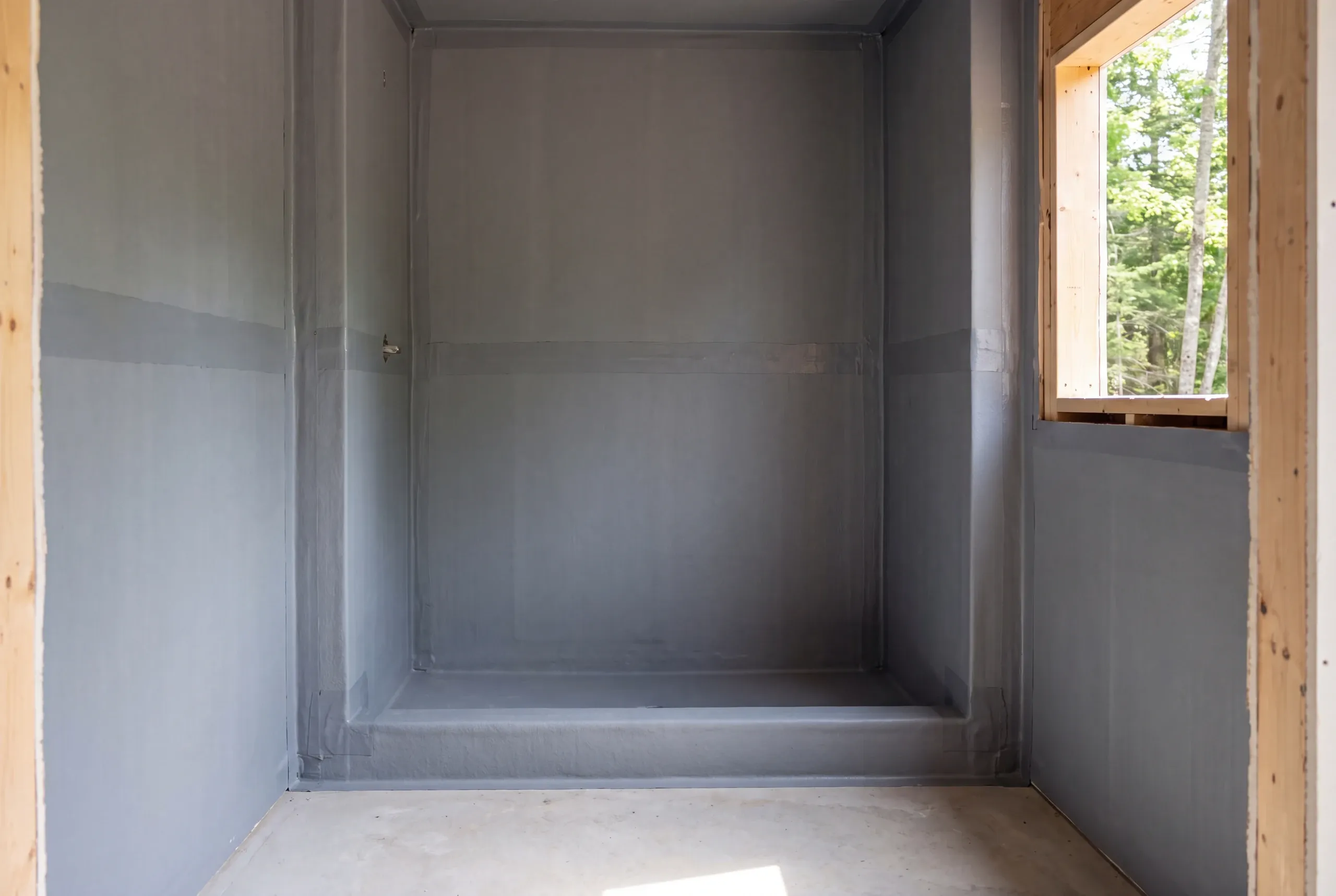 An empty shower alcove coated with a gray continuous waterproofing membrane, showing sealed seams and a window opening to greenery on the right.