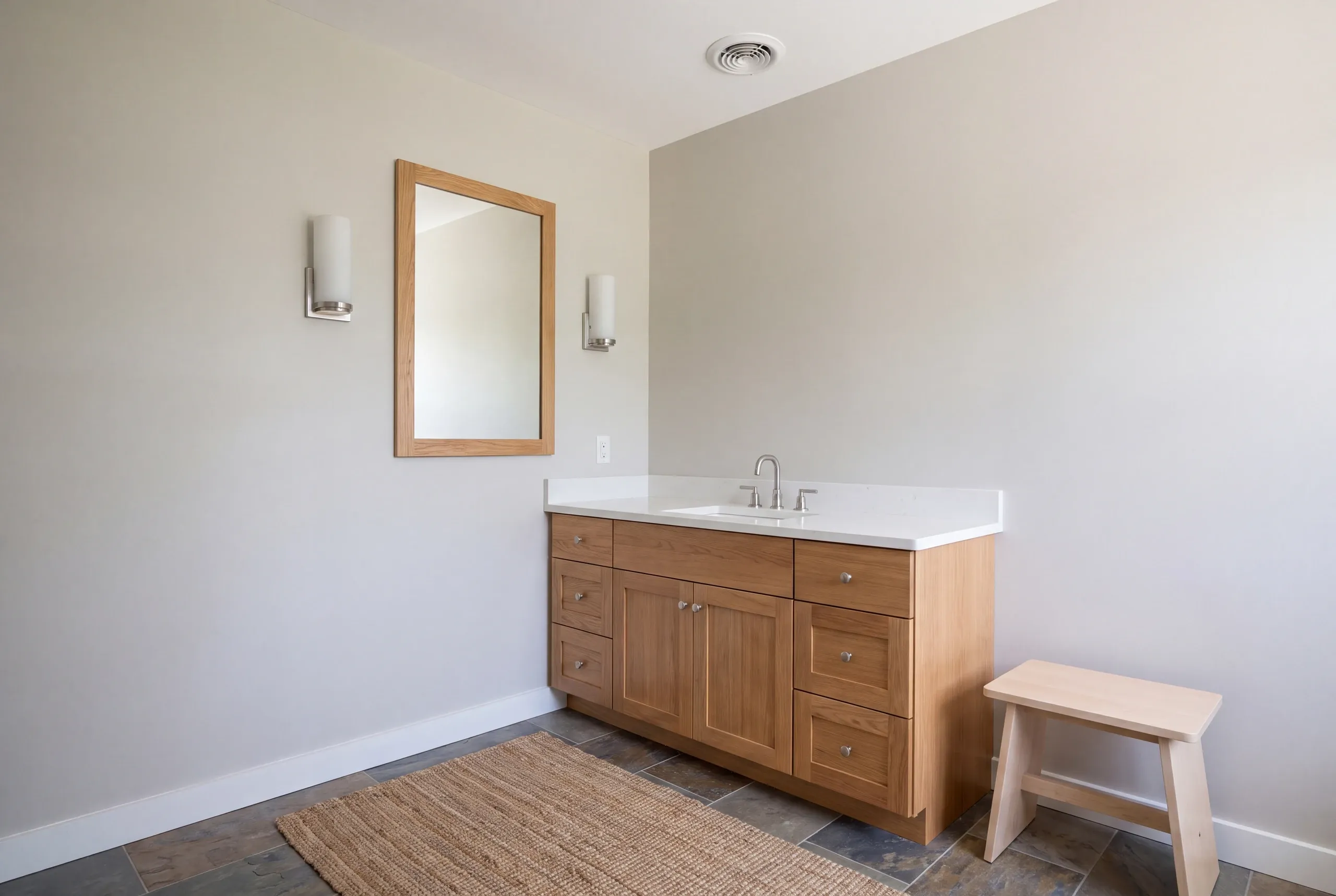 A finished vanity with a wooden cabinet, white countertop, mirror, balanced sconces, and a ceiling exhaust grille.