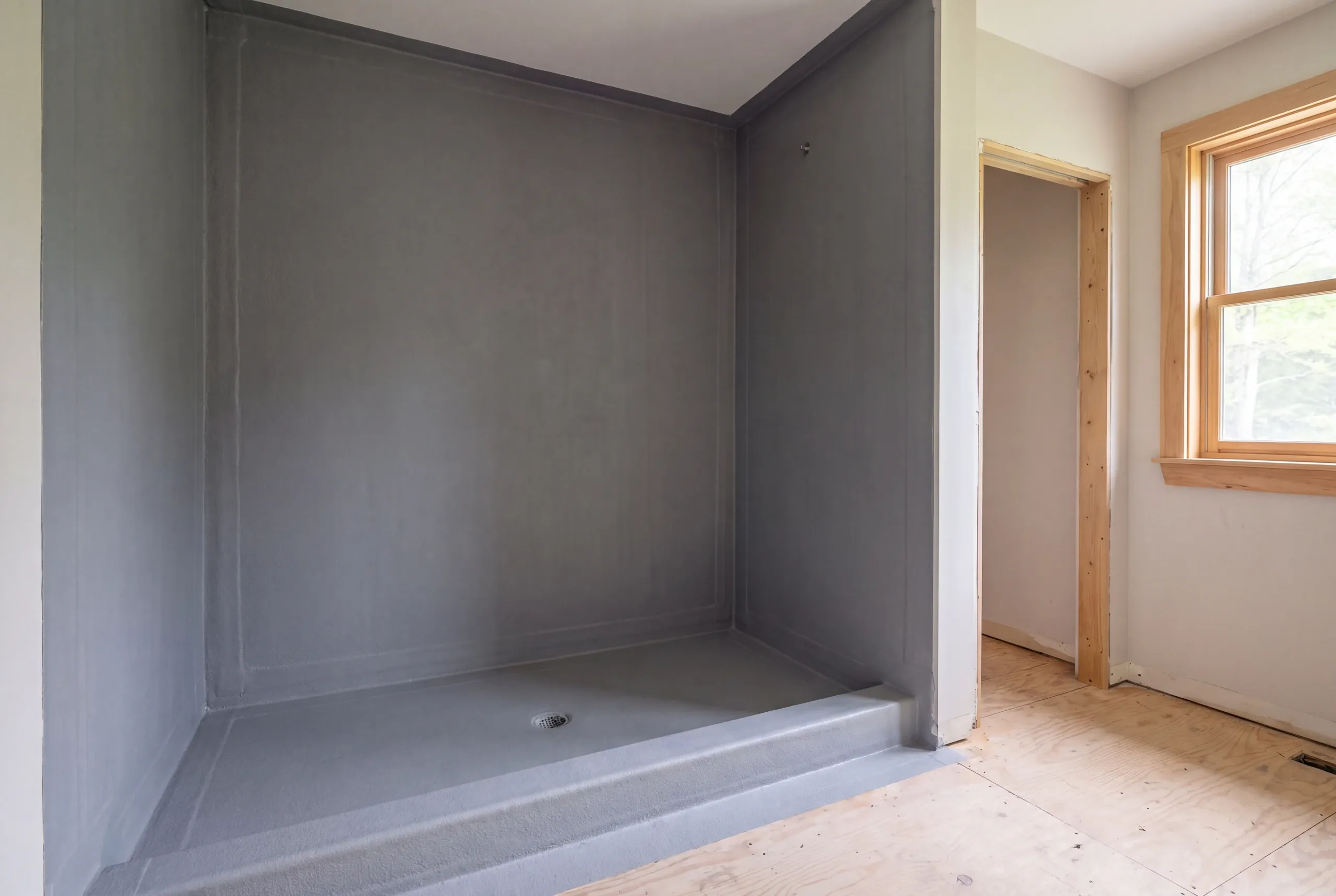 An empty shower alcove coated with grey waterproofing material and a drain, with bare plywood flooring outside.