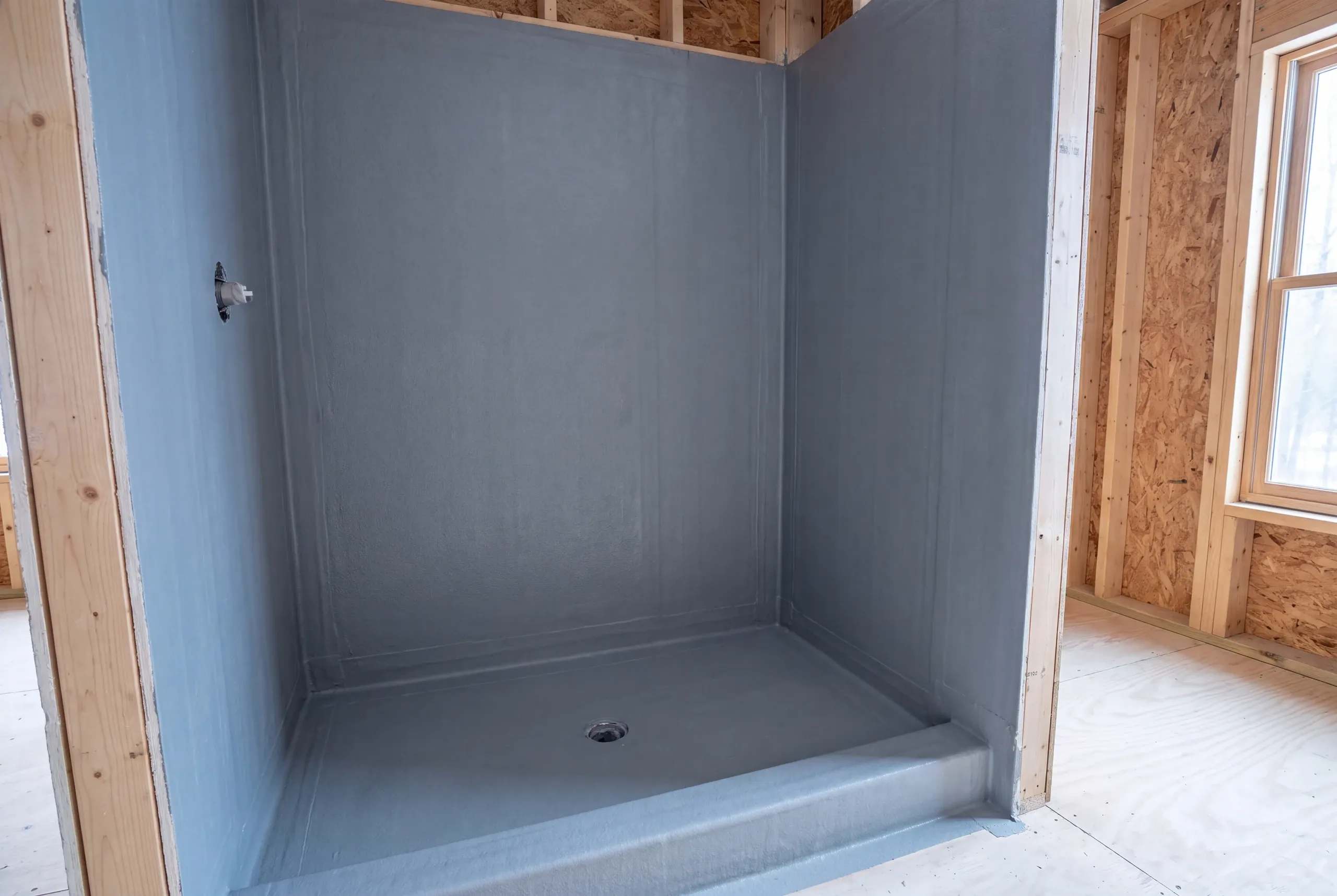 An empty shower alcove with gray waterproofing membrane applied to the walls and pan, surrounded by unfinished wooden framing.