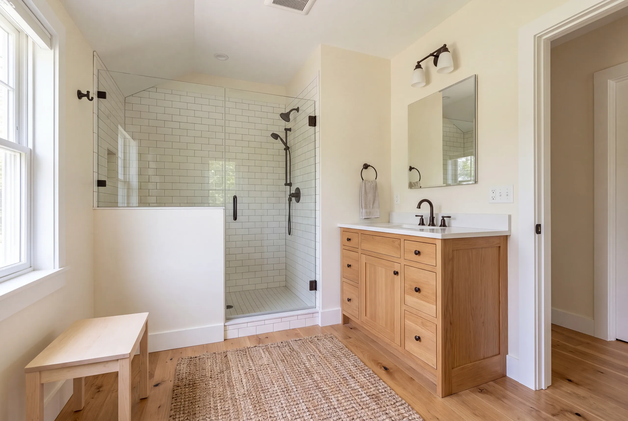A finished bathroom features a glass-enclosed shower with white subway tile, a wooden vanity with a white countertop and mirror, and a small wooden bench.