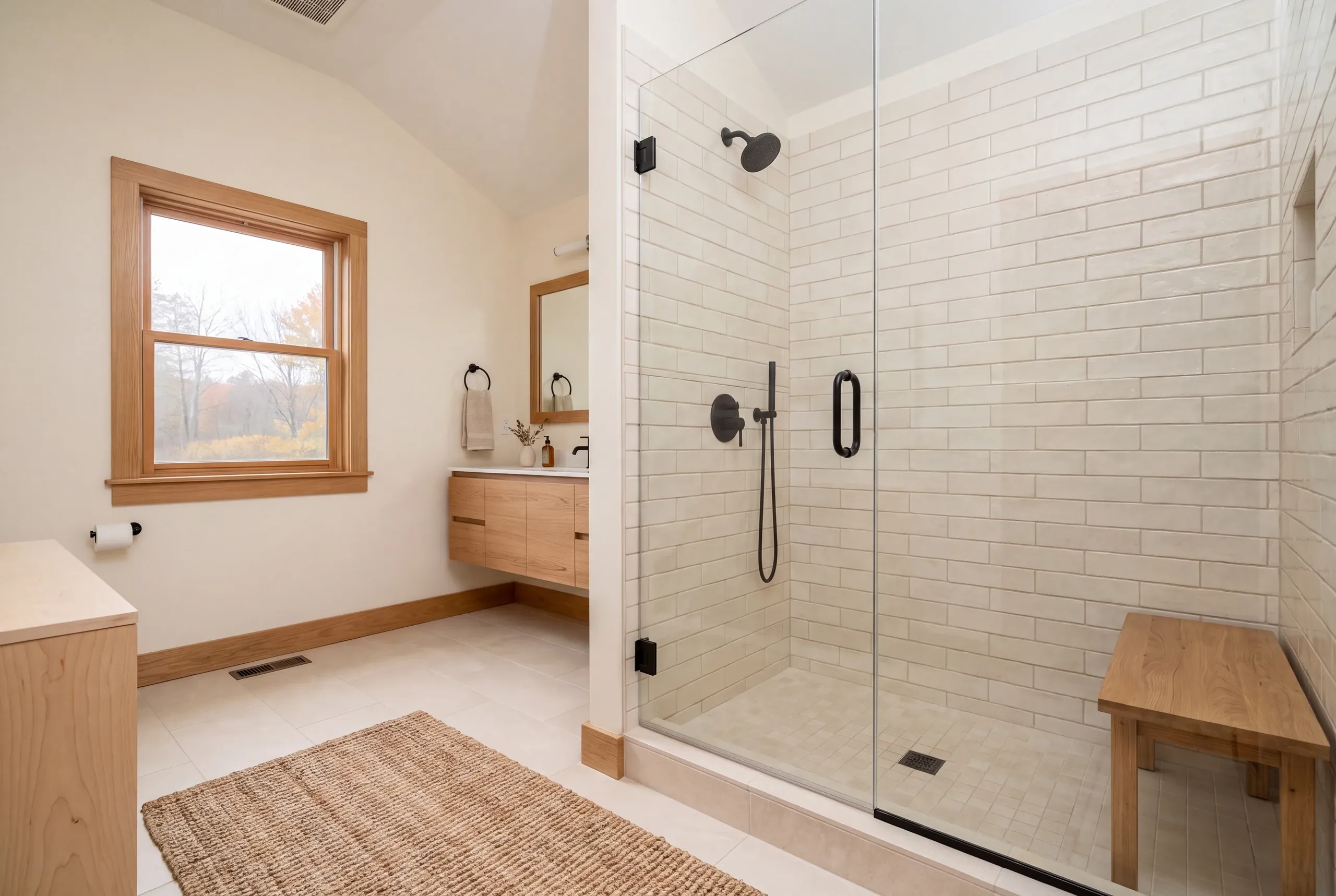 A modern tiled shower enclosure with glass doors and a wooden bench stands inside a bright bathroom with light wood trim.