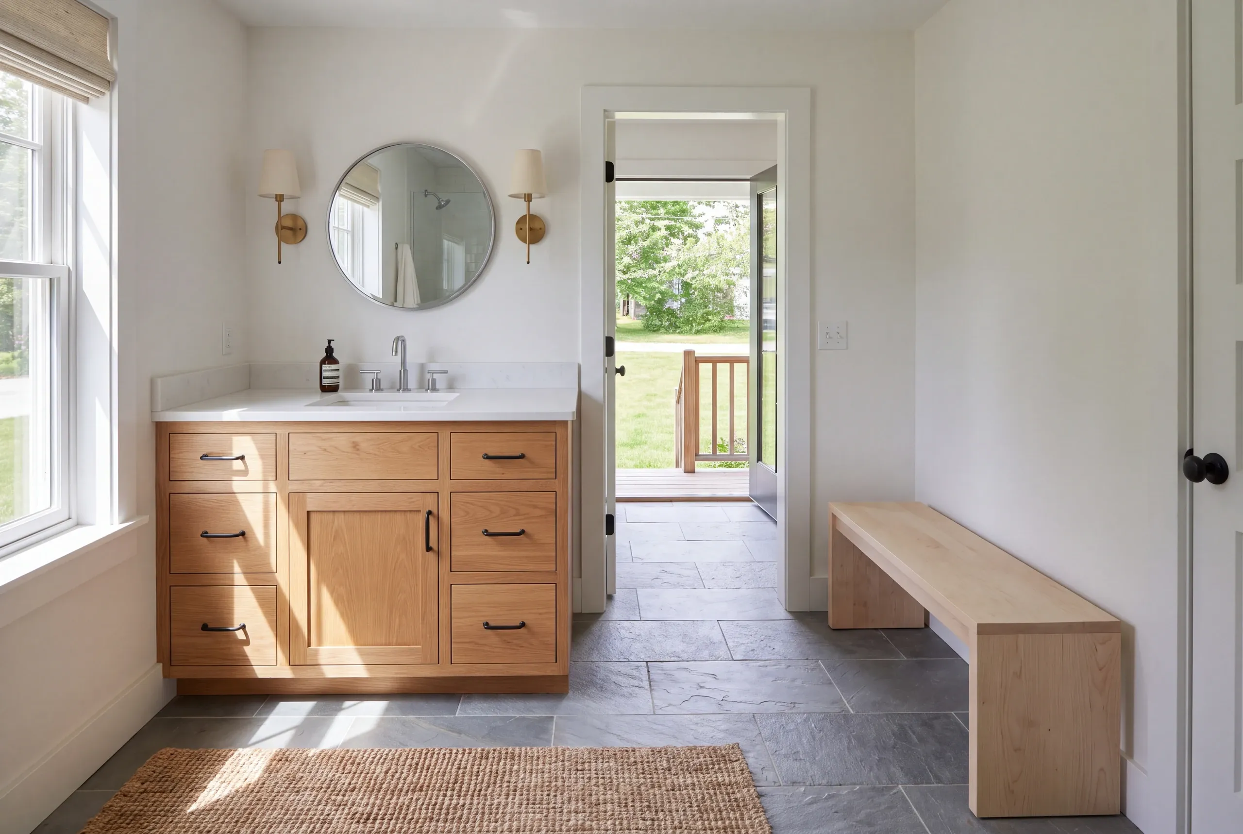 A bright bathroom features a wooden vanity with a white countertop, round mirror, and sconces, alongside a wooden bench and slate tile flooring.