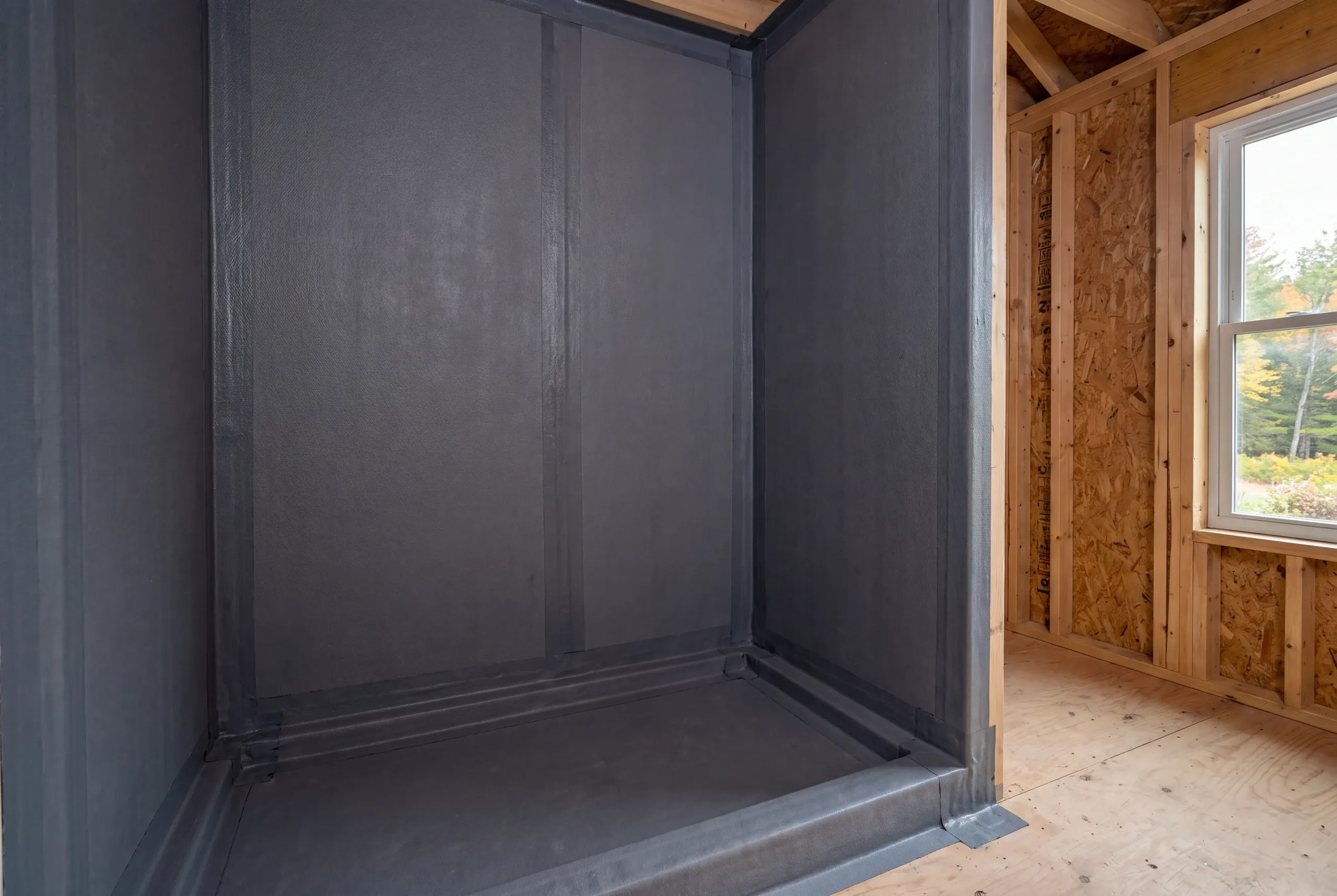 An empty shower alcove covered in a dark gray waterproofing membrane with sealed seams, situated next to unfinished wooden framing and a window.