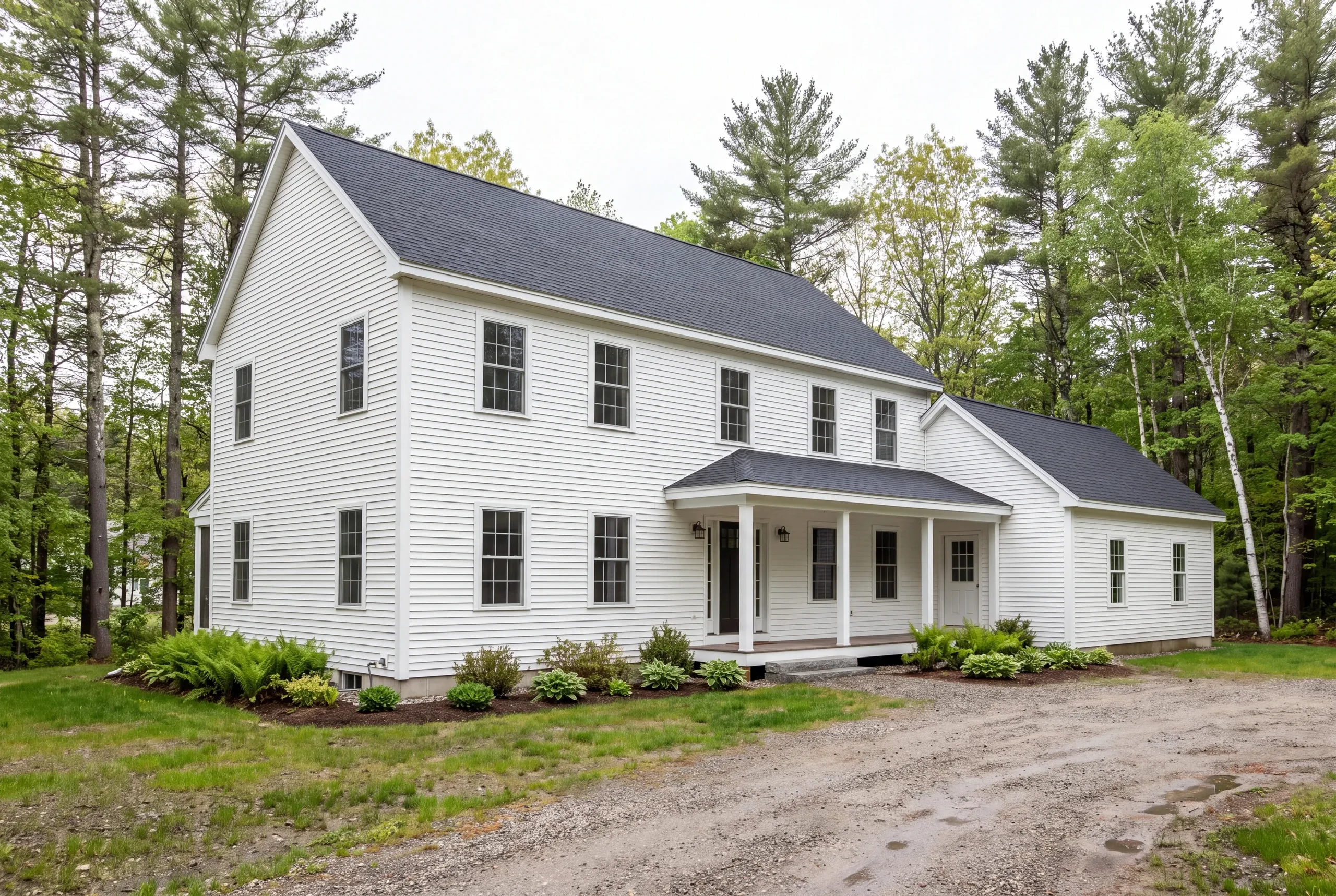 Exterior view of a white two-story New England style home with a dark roof and a front porch, surrounded by trees and a dirt driveway.