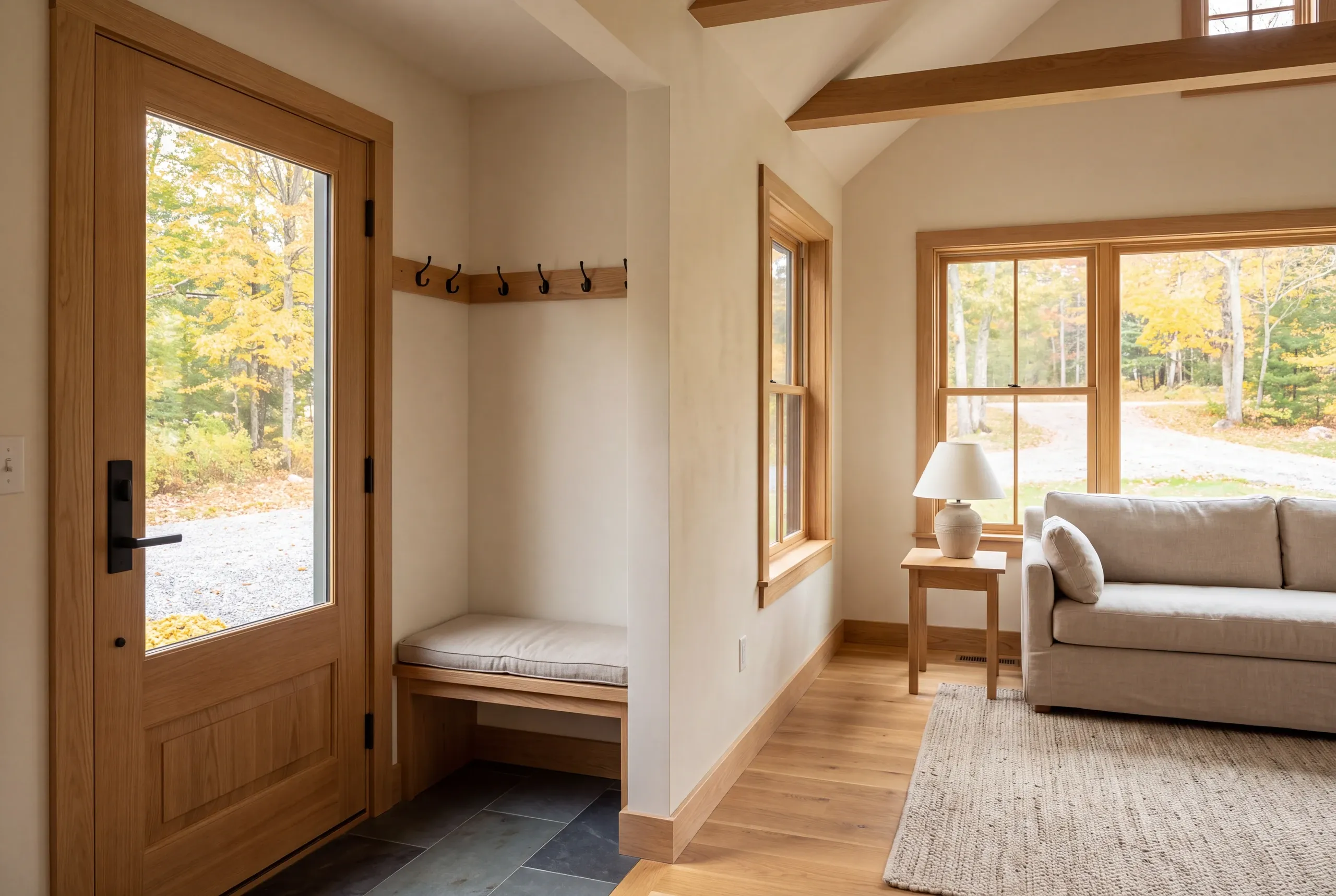An interior shot showing an entryway with a wooden door, bench, and coat hooks connected to a living room with a sofa and windows.