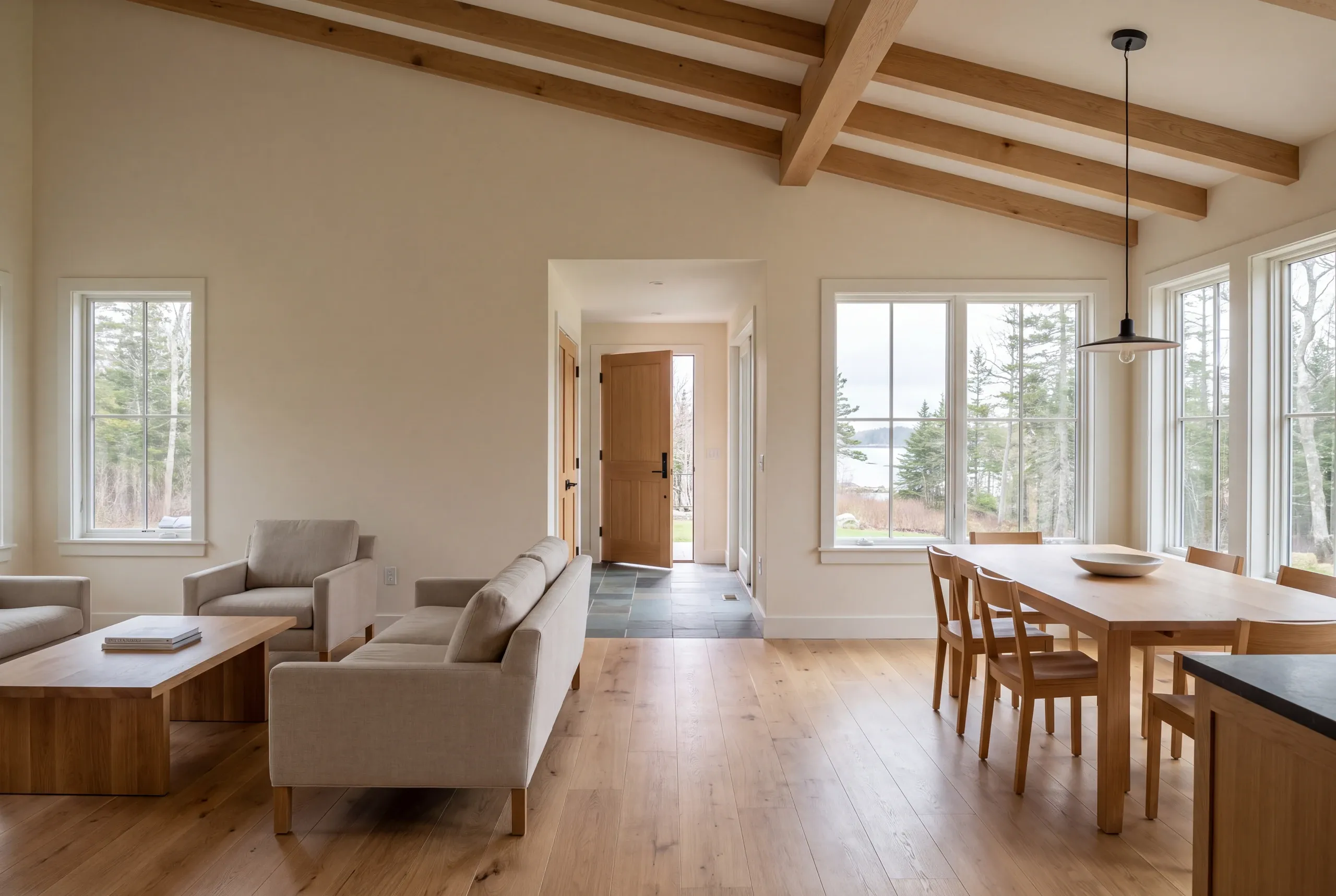 A bright living and dining room featuring exposed wooden ceiling beams, light wood flooring, neutral furniture, and large windows looking out to a wooded view.
