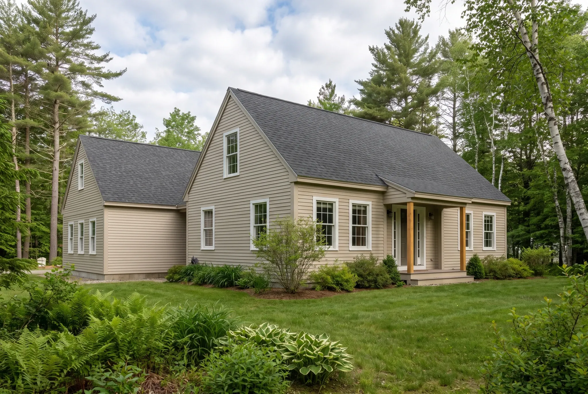 General contractor in Bowdoin, ME: exterior view of a beige clapboard house with dark shingled roofs and a small front porch surrounded by trees and a green lawn.