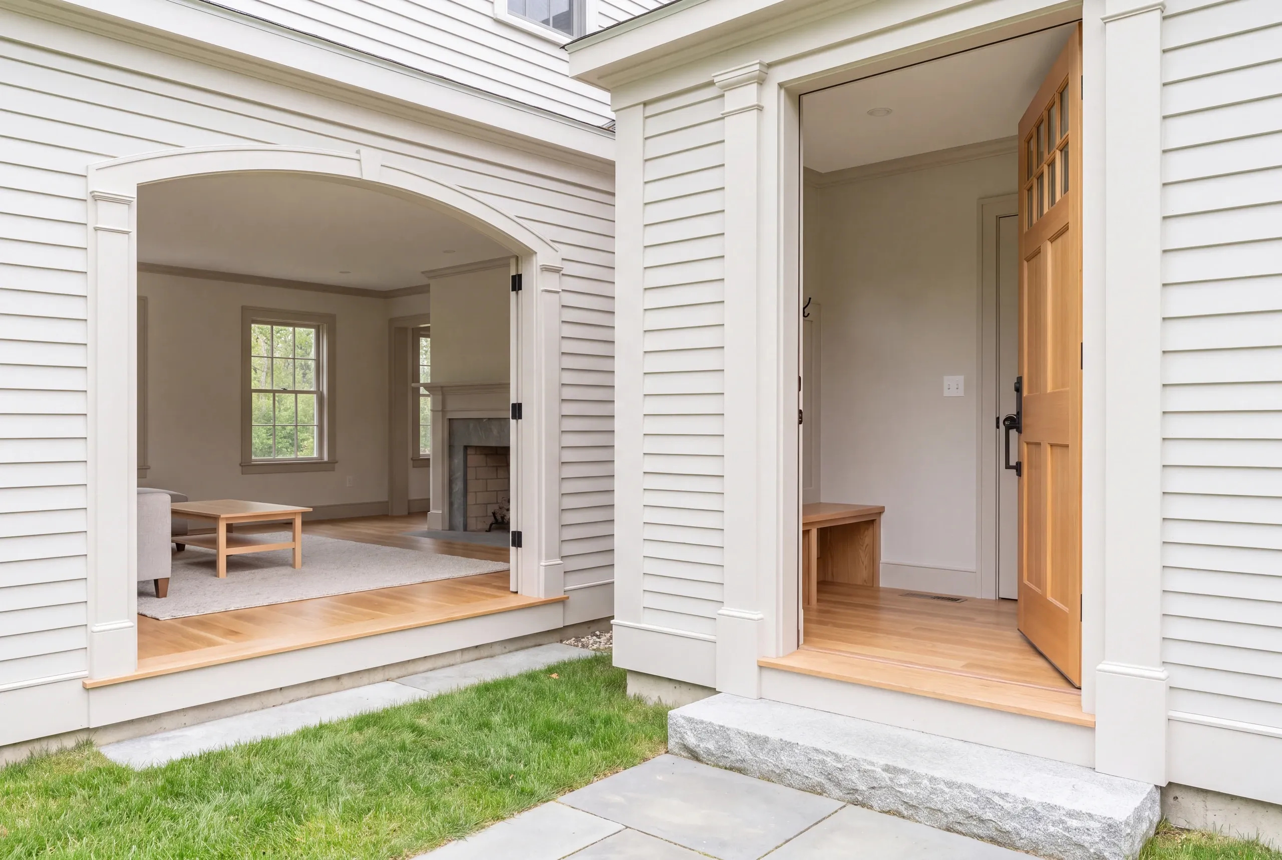 Exterior view showing white clapboard siding, a stone patio, an open arched opening leading to a furnished living room, and an open wooden door leading to an entryway with a bench.