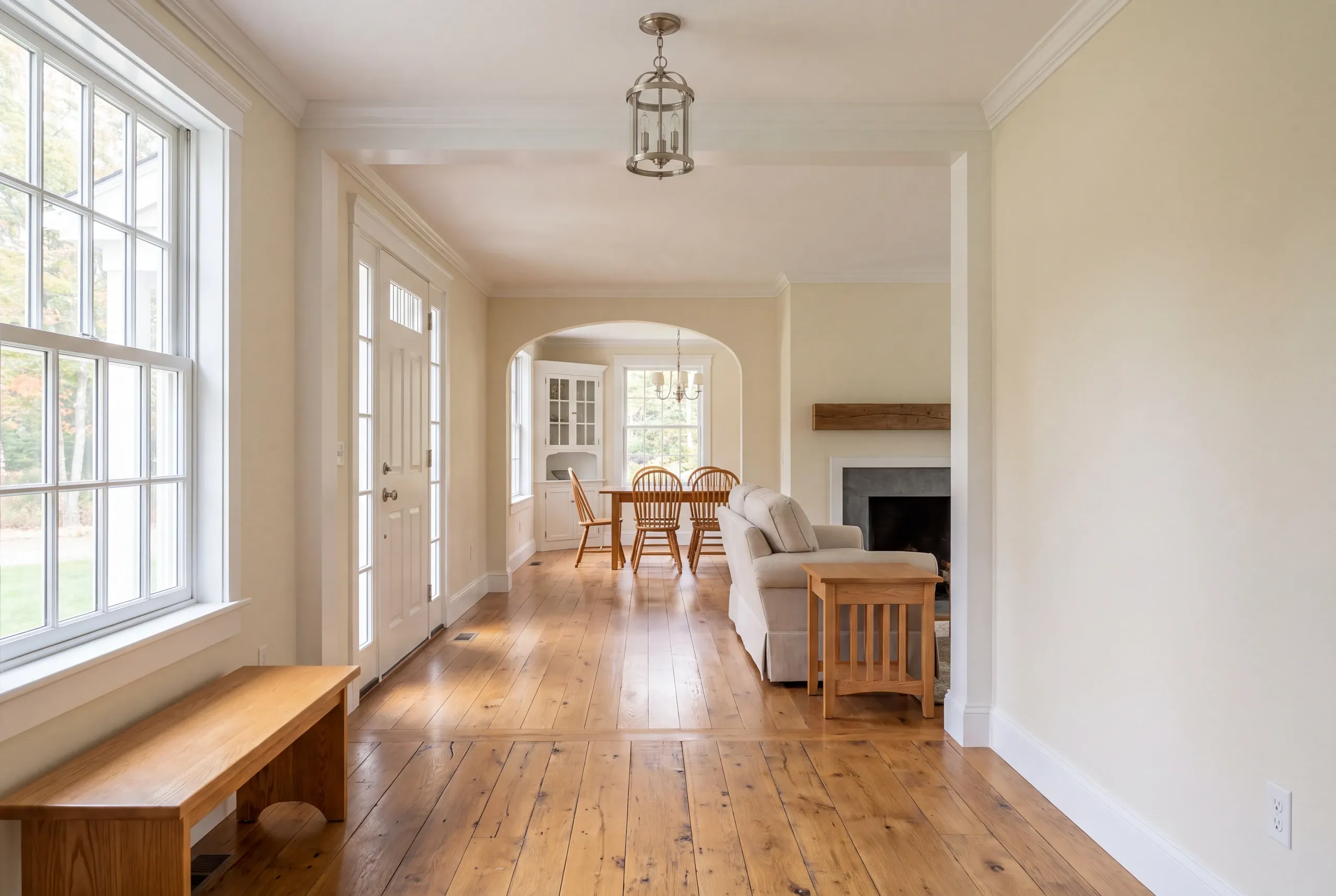 Interior view showing a hardwood floor hallway connecting to an entryway, a living room with a sofa and fireplace, and a dining area in the background.