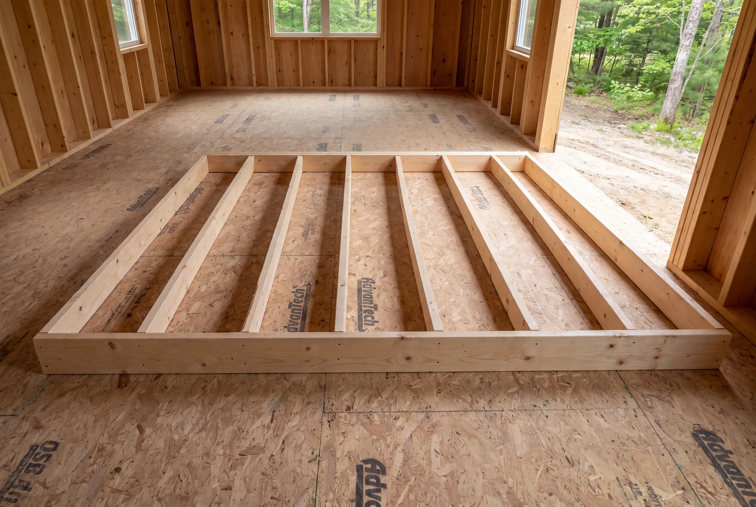 A wooden interior wall frame lies flat on a subfloor inside a room under construction with visible wall studs and windows.