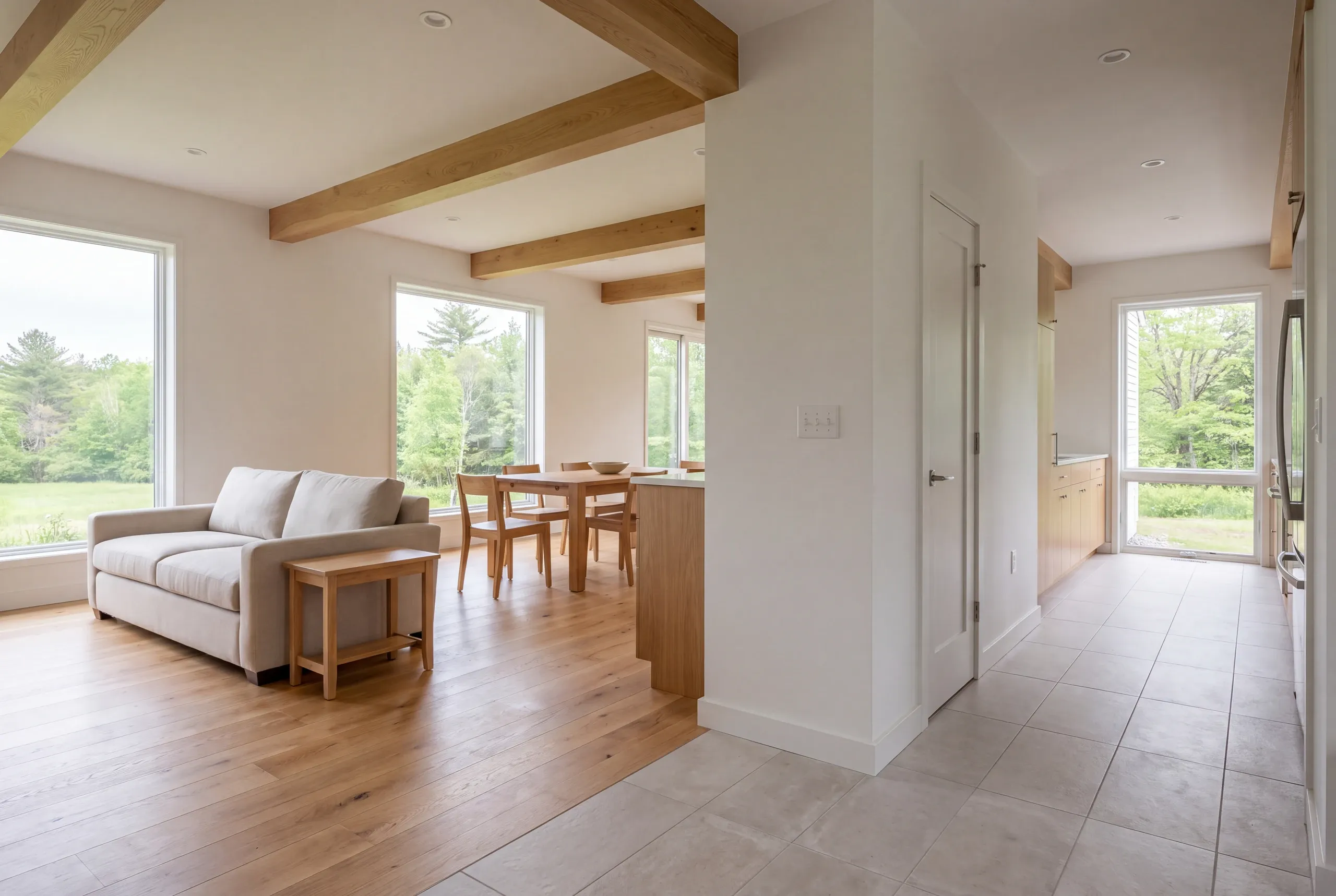 Interior view of a living room with a beige sofa, wooden beams on the ceiling, and an adjoining dining and kitchen area with tiled flooring.