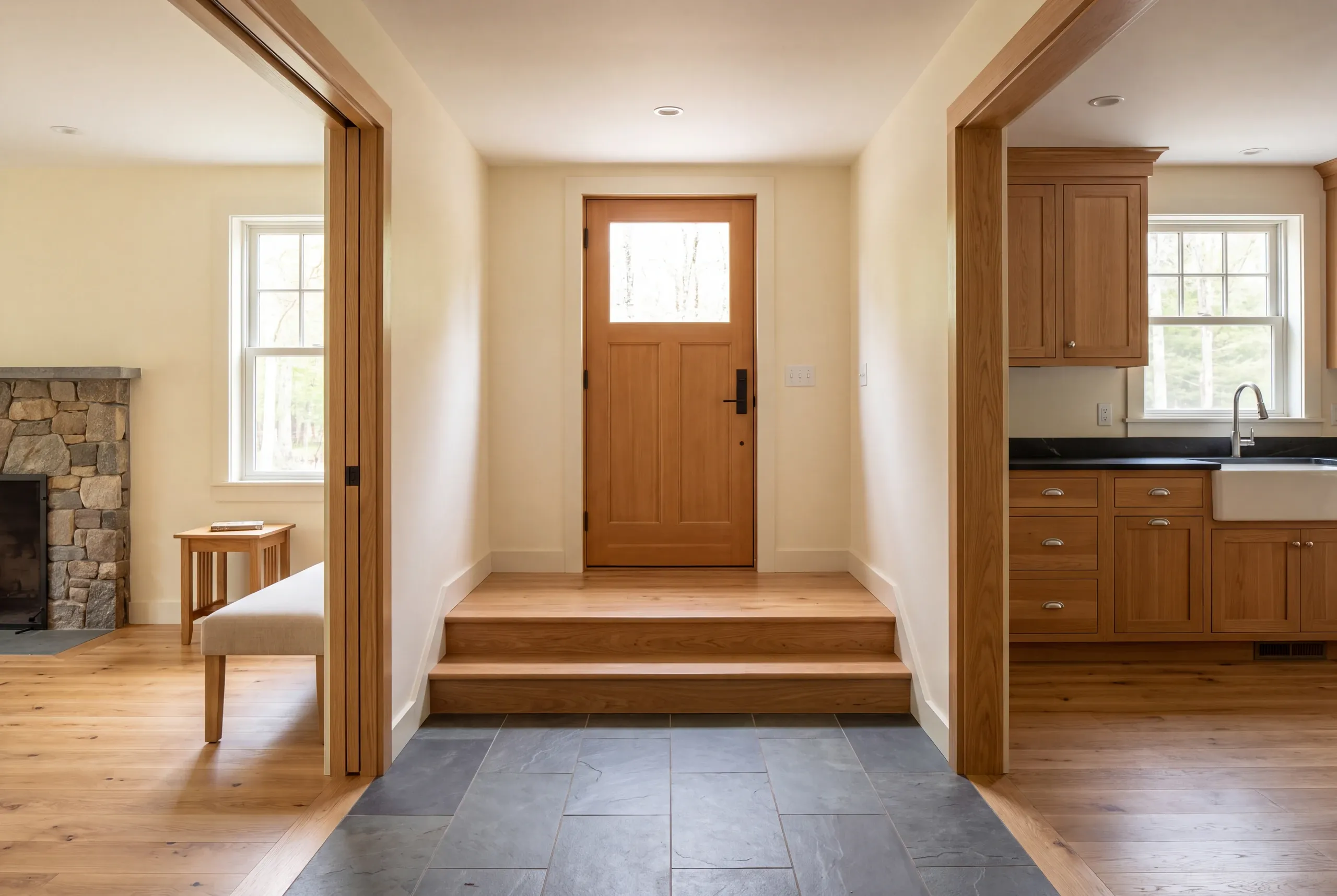 A finished indoor entryway features dark stone floor tiles transitioning to wood steps leading up to a wooden front door, flanked by a living area on the left and a kitchen on the right.