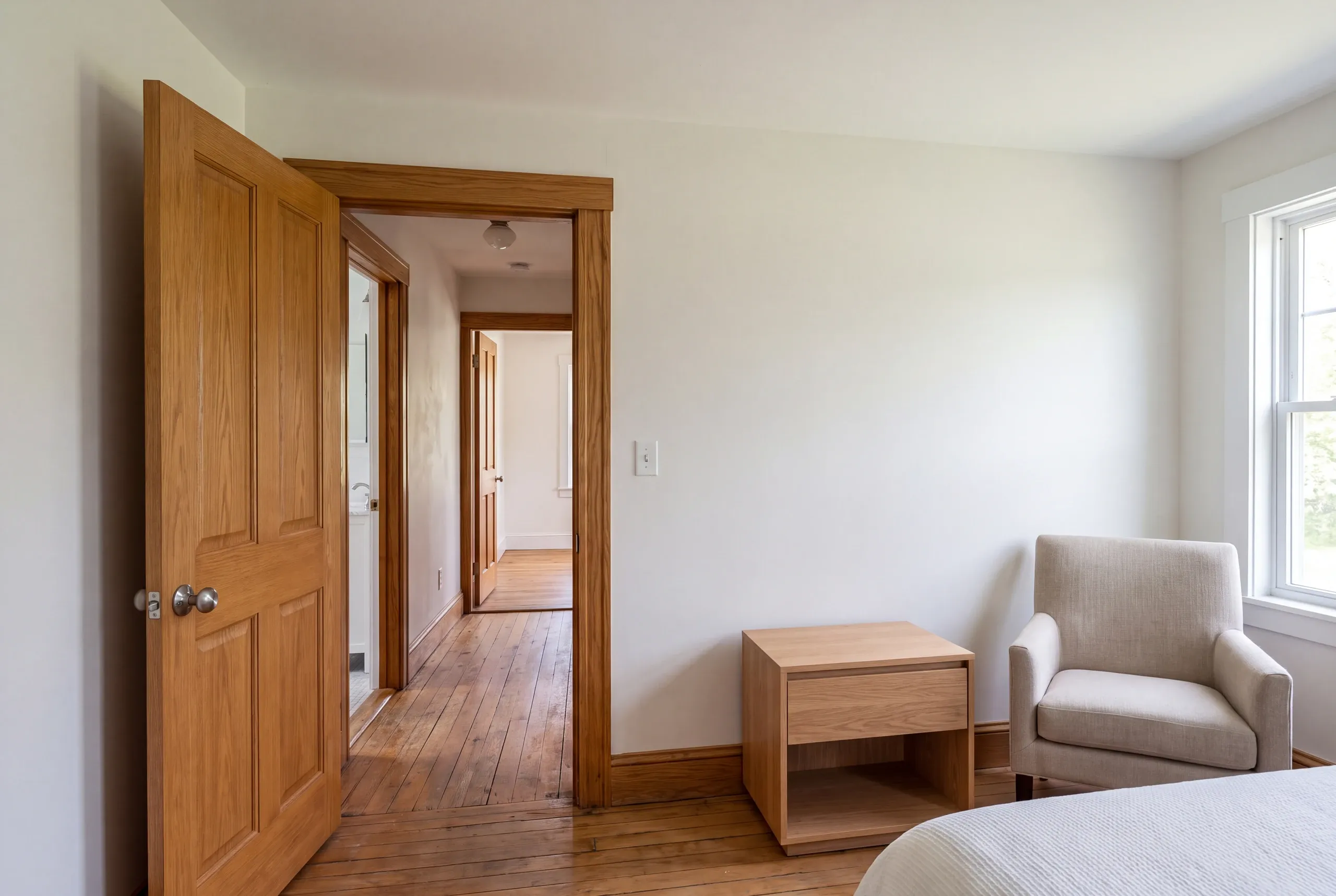 A bright finished bedroom featuring a wooden bedside table, an armchair, and an open doorway revealing a wooden hallway and a bathroom.
