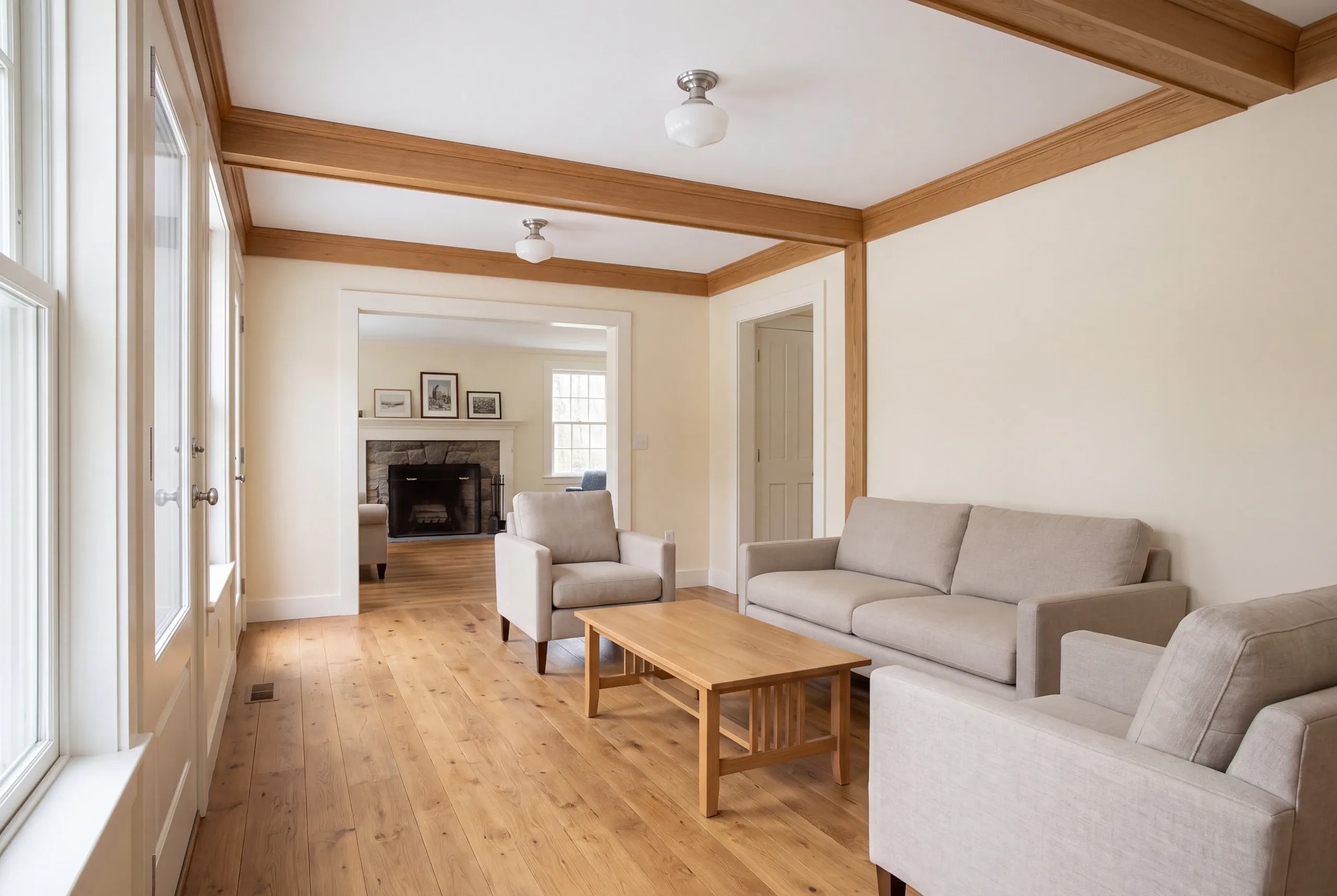 A finished family room with hardwood floors, wooden ceiling beams, and neutral-toned seating looking into an adjoining room with a stone fireplace.