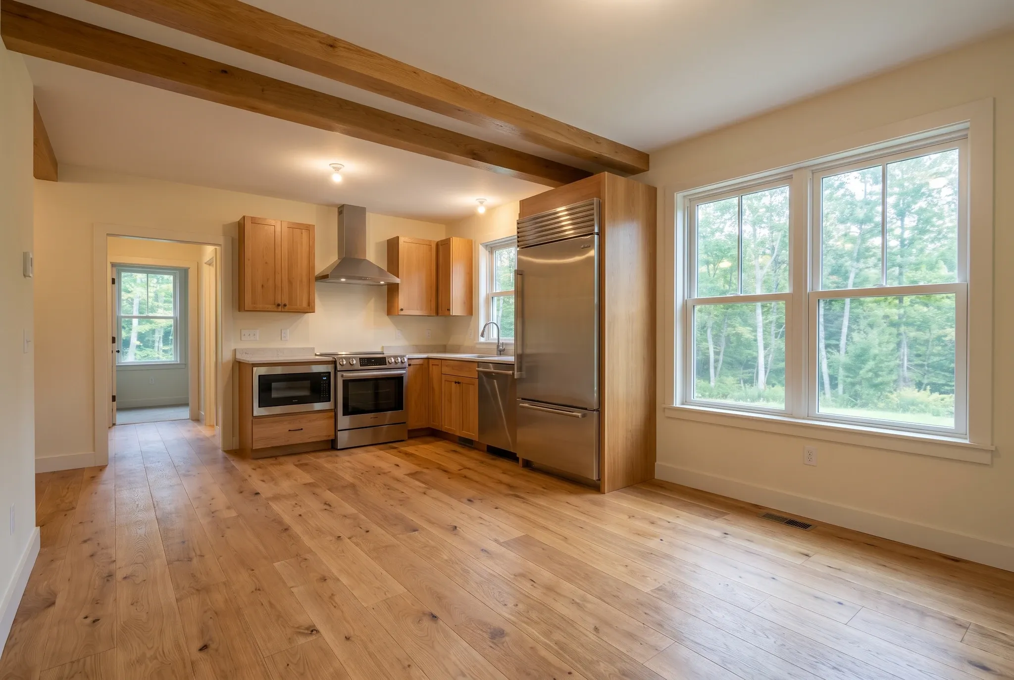 Interior view of a finished kitchen featuring wooden flooring, cabinets, stainless steel appliances, and large windows looking out onto trees.