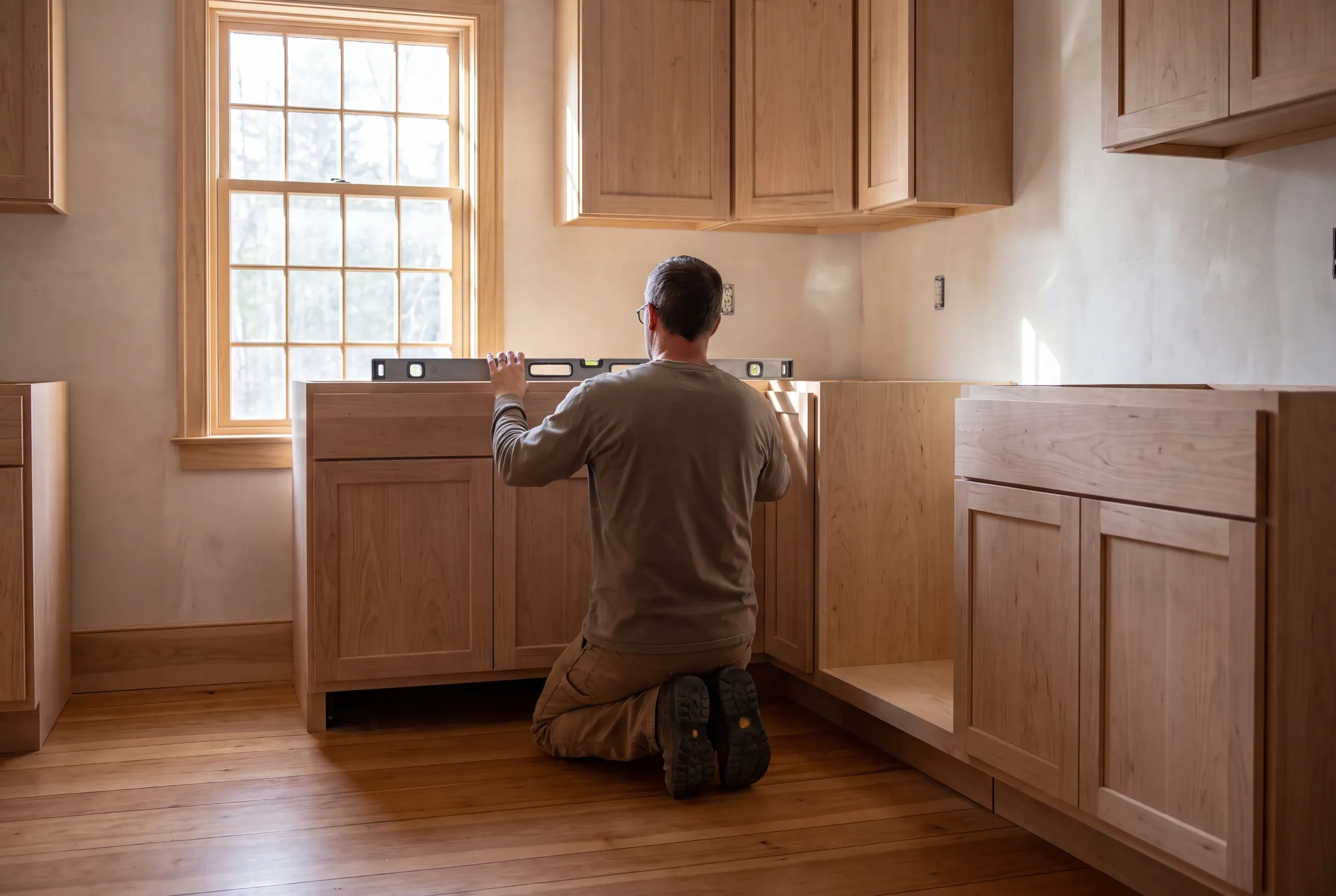 An installer kneels on a wooden floor in a kitchen, using a spirit level to align base cabinets below upper cabinets.
