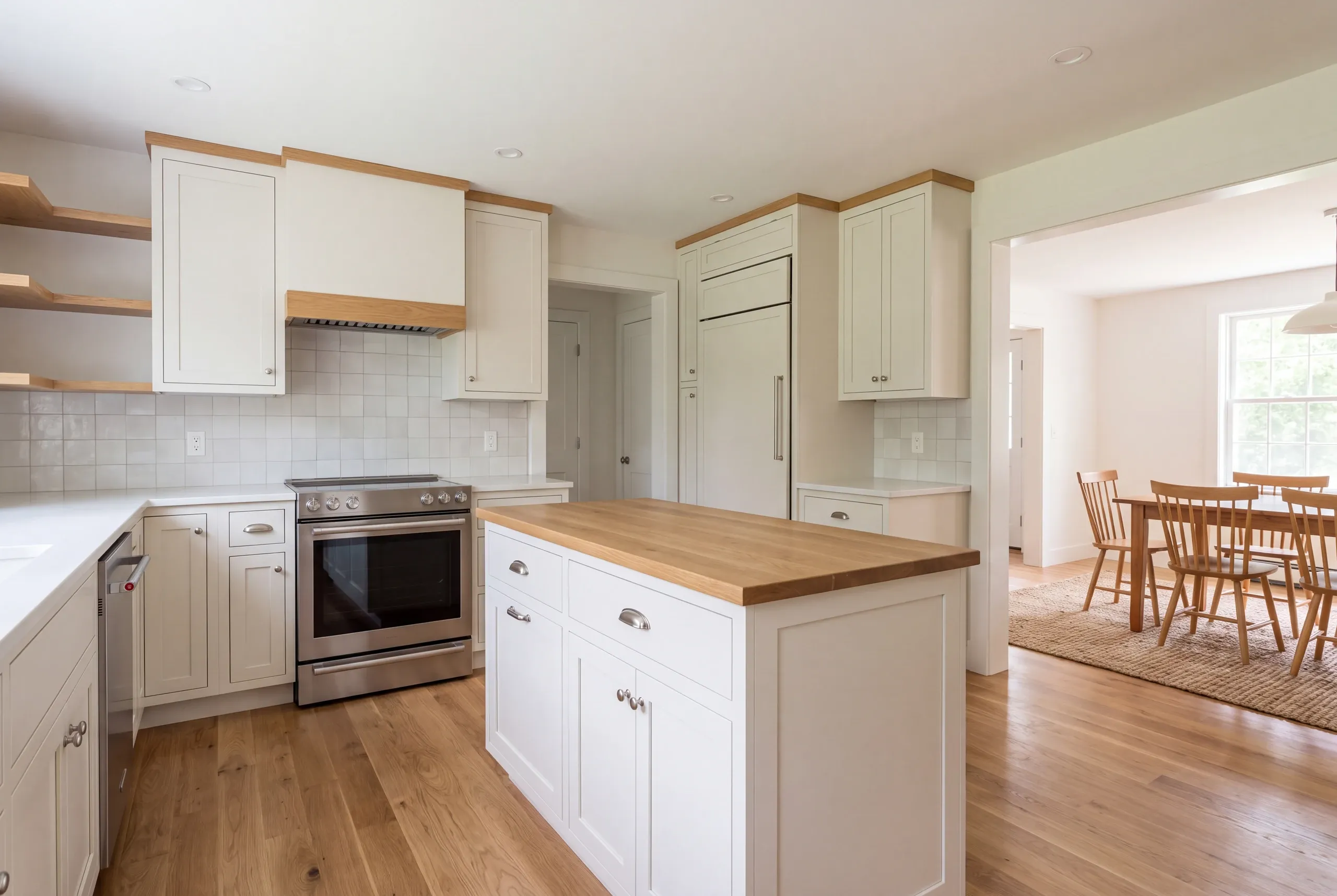 A newly finished white kitchen features a central wooden-topped island, stainless steel appliances, open shelving, and an open entryway leading to an adjacent dining area.